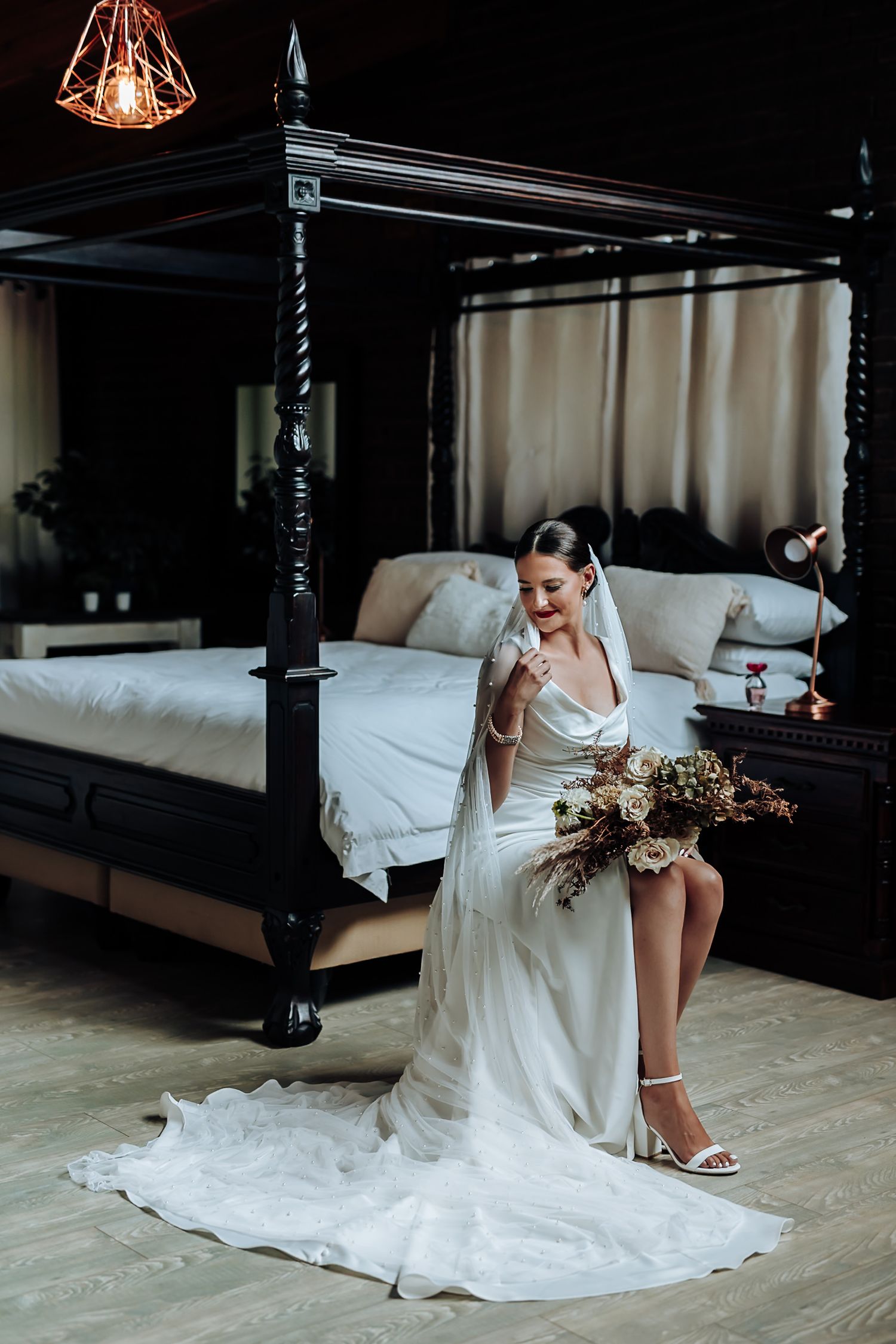 Elegant bridal scene with flowing white gown, dried floral bouquet and antique four-poster bed in moody lighting.