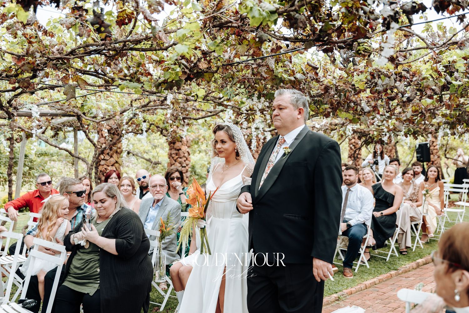 Outdoor wedding ceremony under grape vine covered arbor with guests seated.