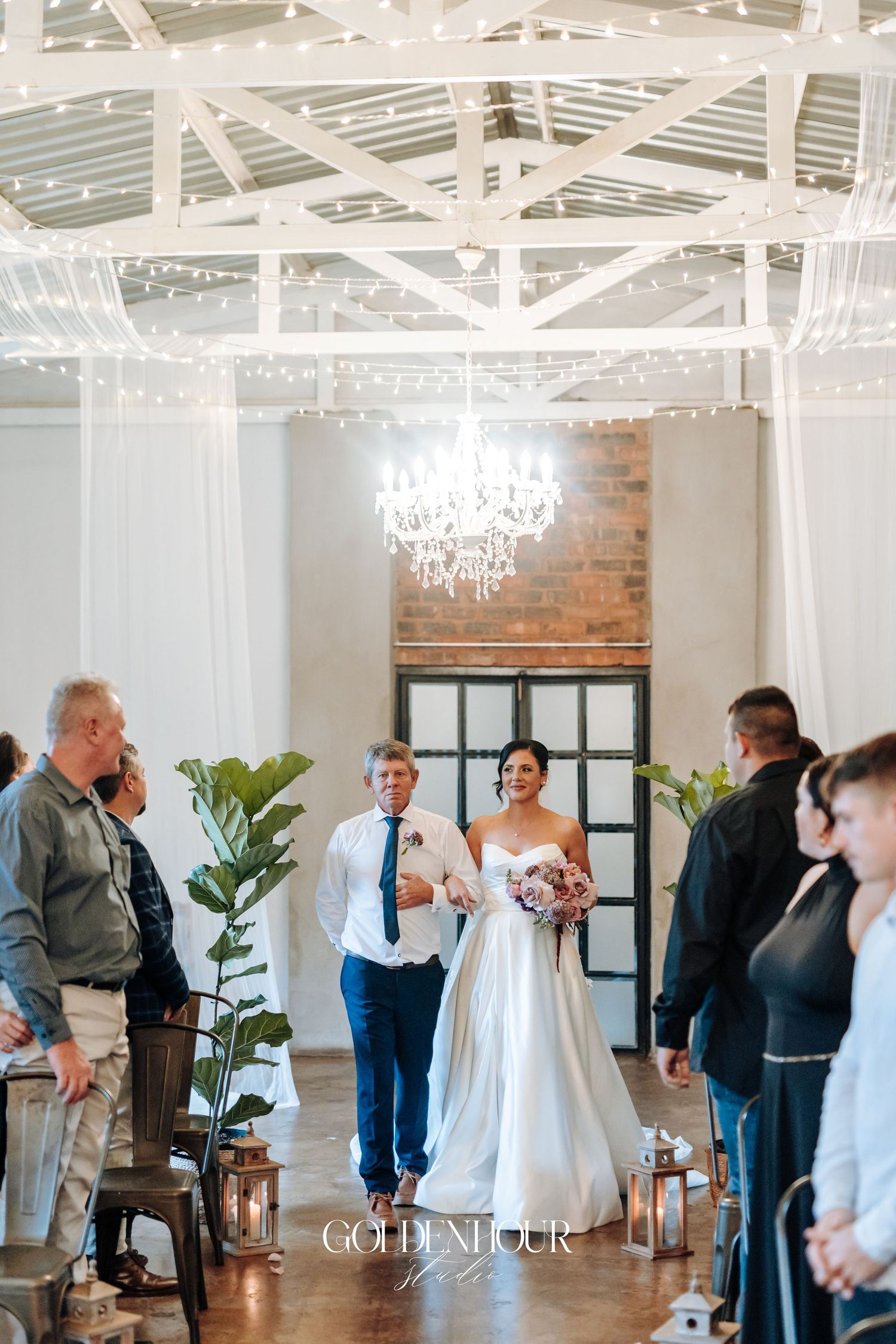 Bride walking down the aisle with escort in a chandelier-lit wedding venue with fairy lights.