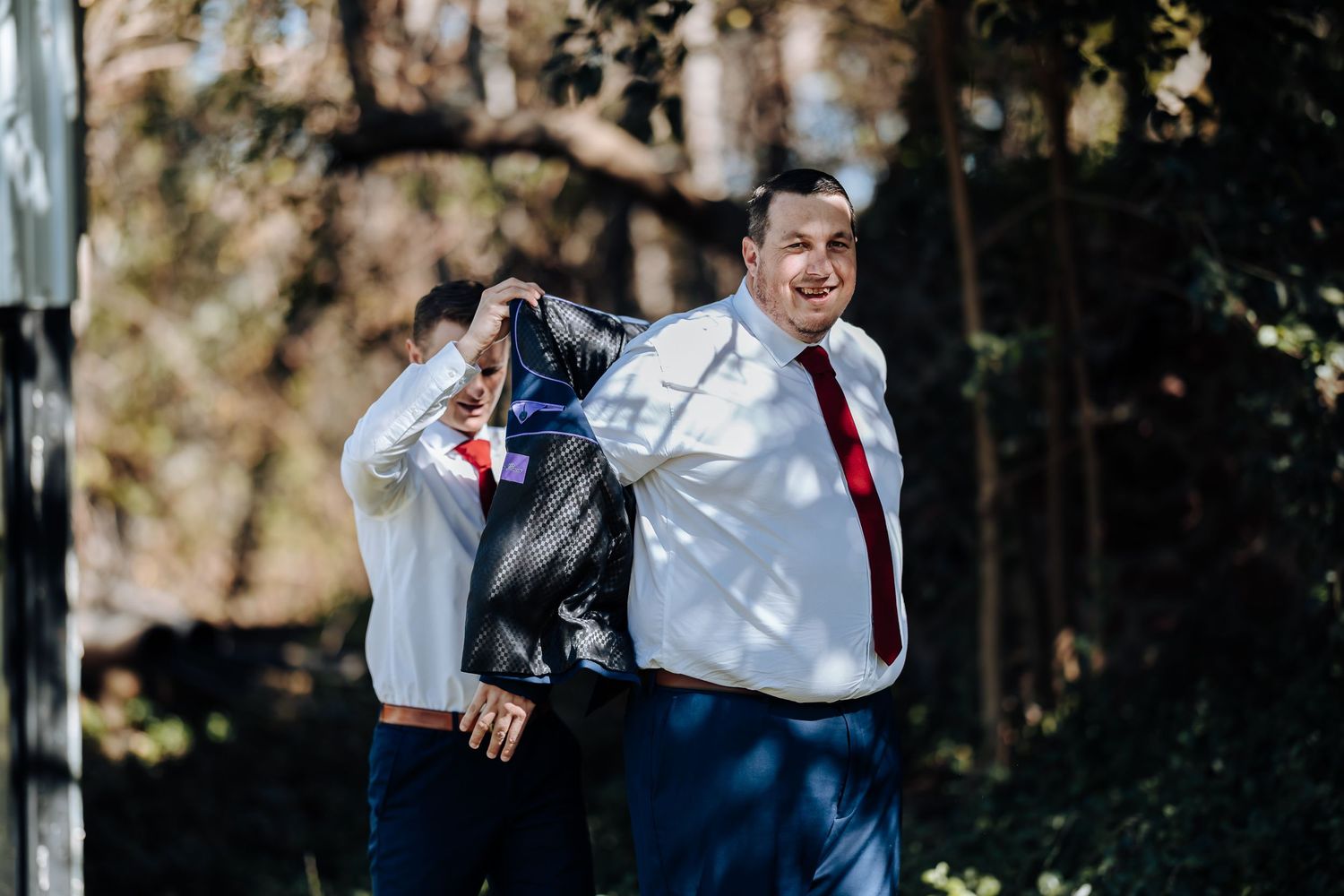 Two groomsmen in white shirts and red ties assist each other with suit jackets outdoors at a wedding.