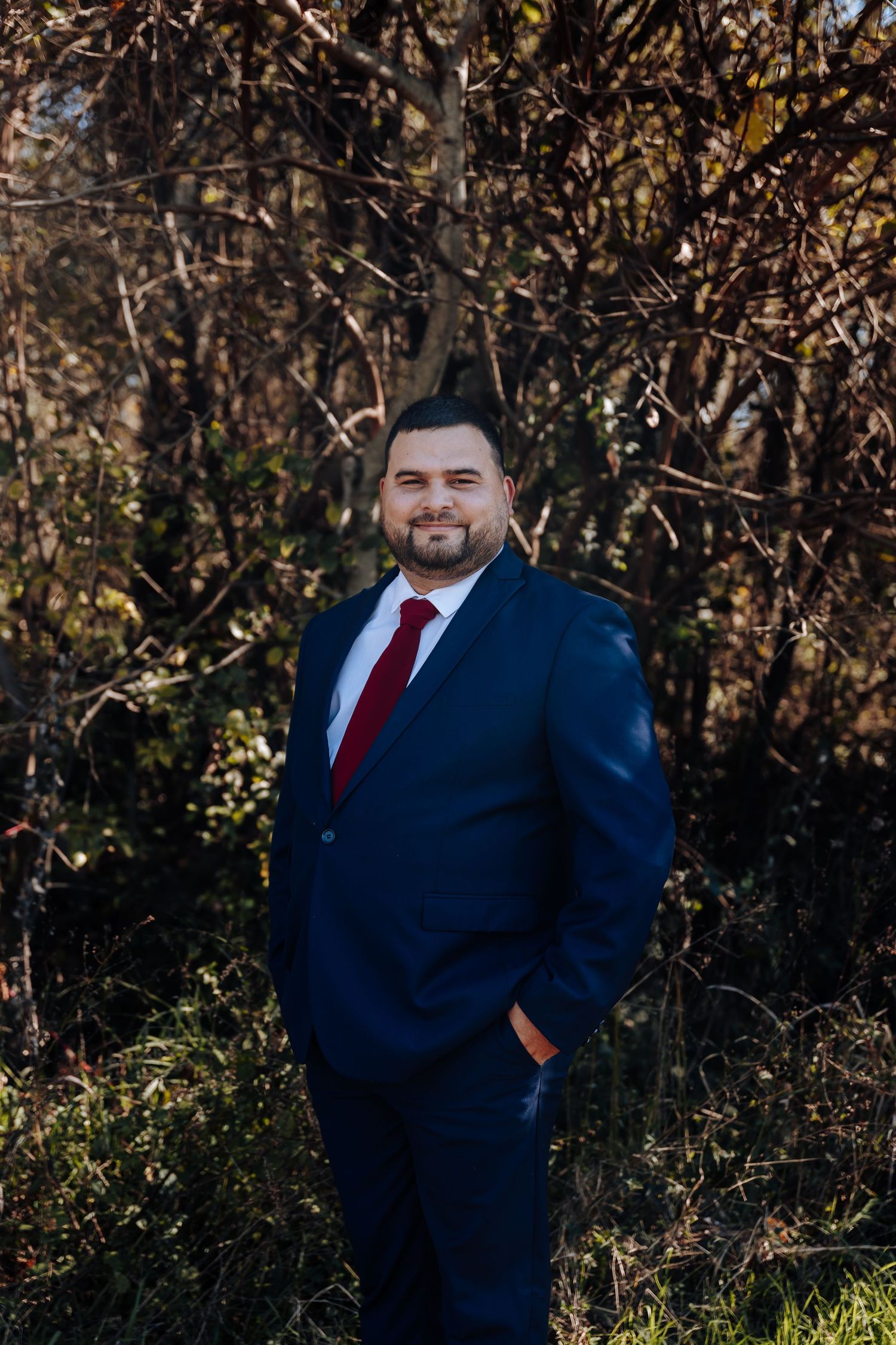 Man in navy suit and red tie smiling outdoors among autumn branches in natural light.
