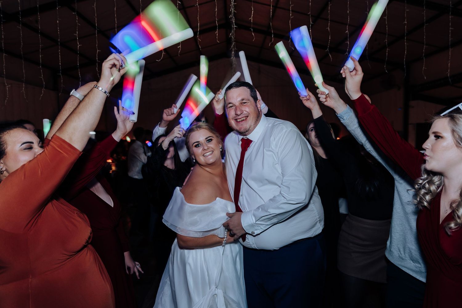Bride and groom celebrate with guests holding colorful LED glow sticks at a festive wedding reception dance.
