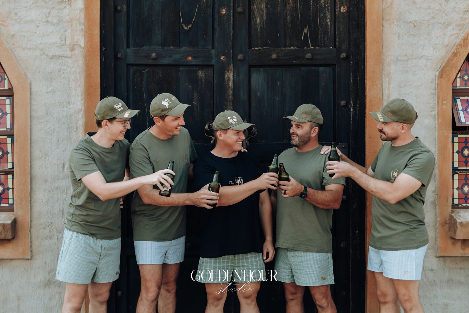 Five men in matching olive green shirts and caps toasting drinks outside a rustic stone building.