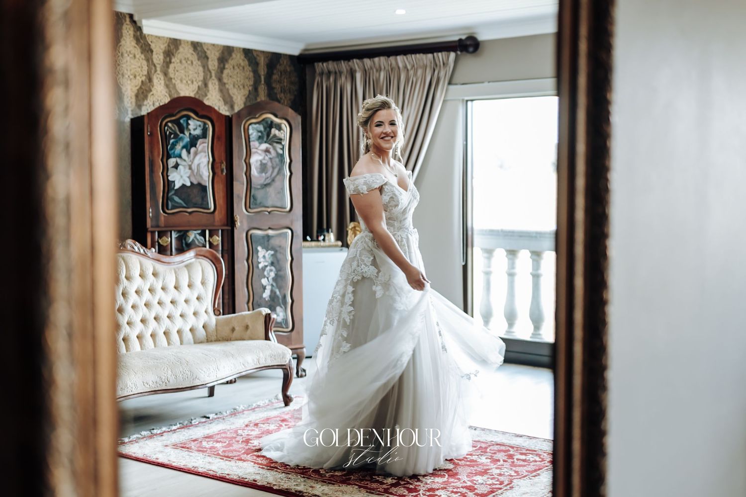 Bride in flowing off-shoulder lace gown posing in ornate vintage-styled bridal suite.