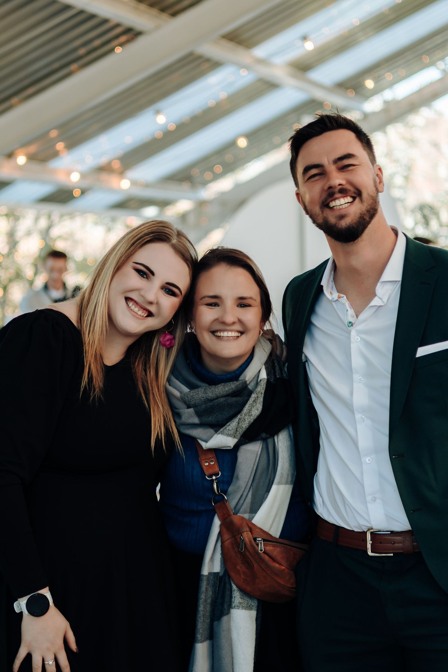Three smiling friends posing together at an indoor event, two women and a man dressed in smart casual attire.
