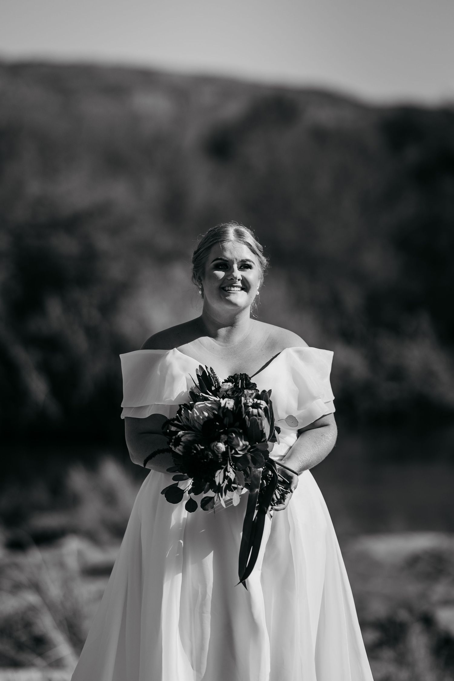 Black and white portrait of a smiling bride in an off-shoulder gown holding a bouquet outdoors.