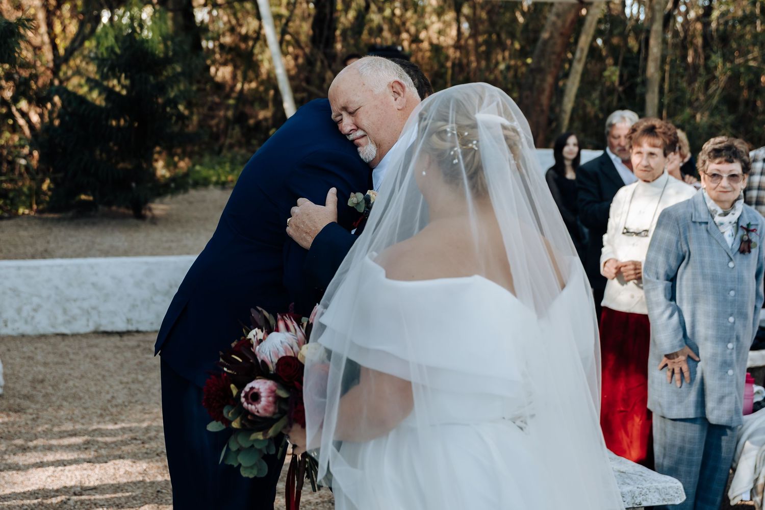 Bride in white gown and veil embracing an older man in navy suit at outdoor wedding ceremony.