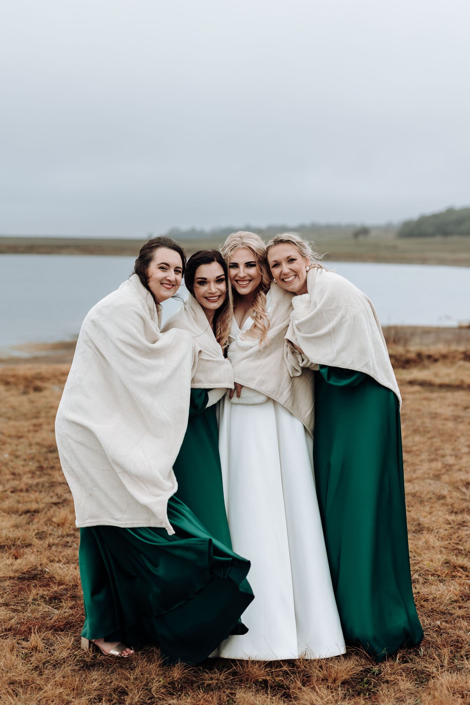 Bridesmaids in emerald dresses huddle together under white blankets by the water.