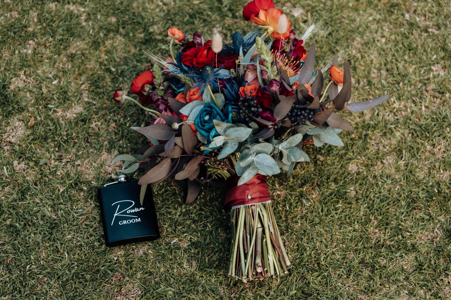 Red and blue wedding bouquet with rustic ribbons displayed on grass.