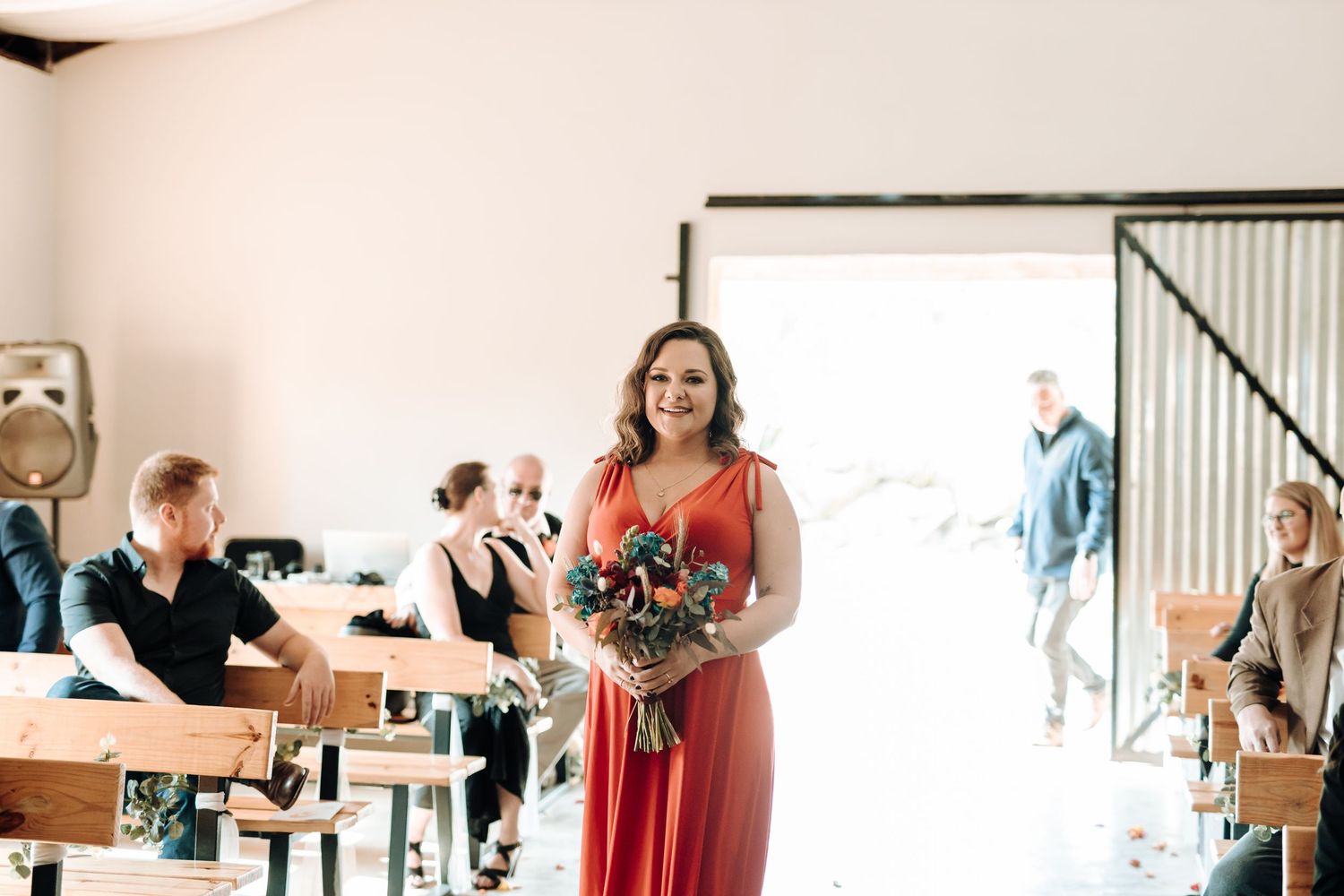 Bridesmaid in coral dress carries bouquet during indoor ceremony processional.