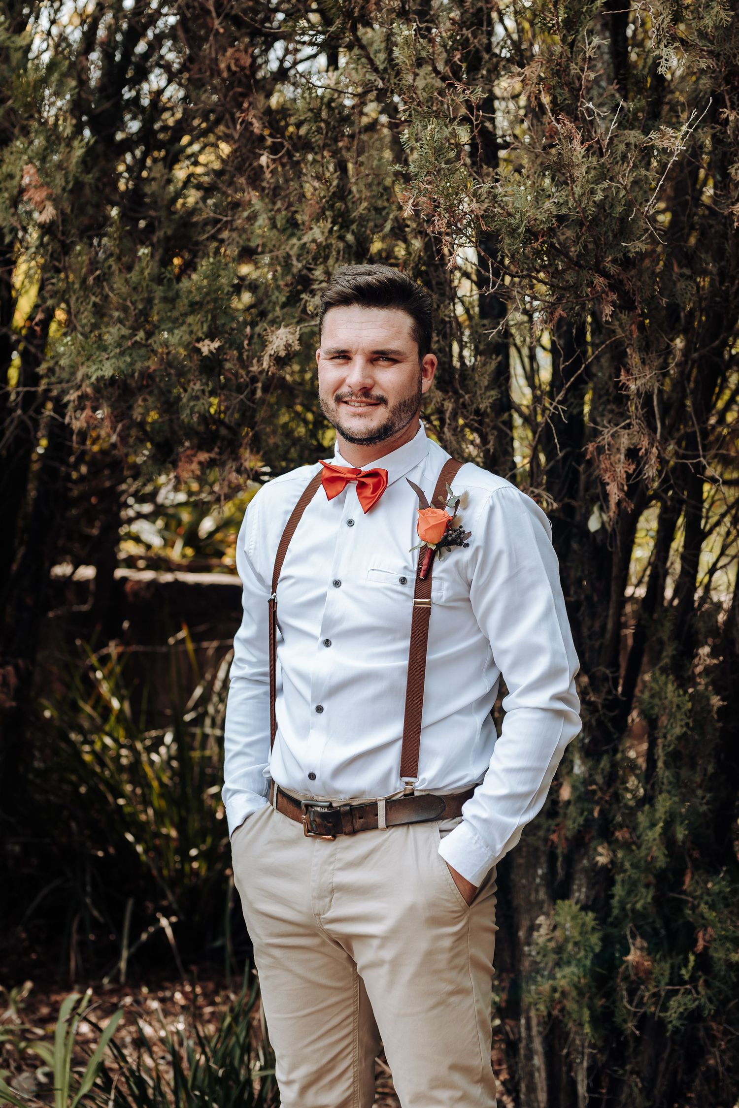 Groomsman in white shirt, brown suspenders and red bow tie poses outdoors.