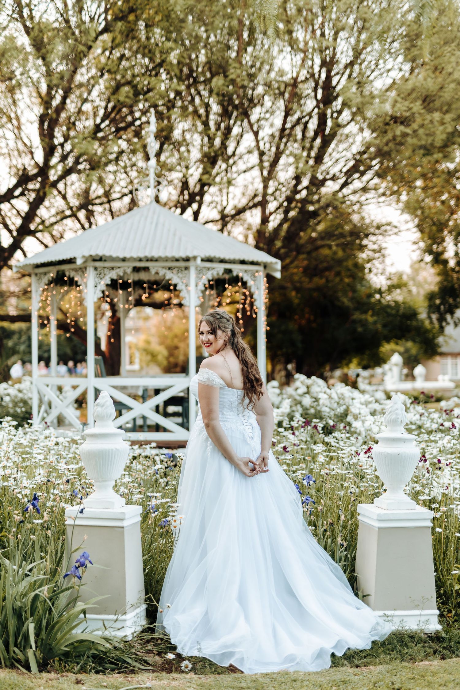 Flowing white wedding gown captured near decorative gazebo amidst blooming wildflower garden.