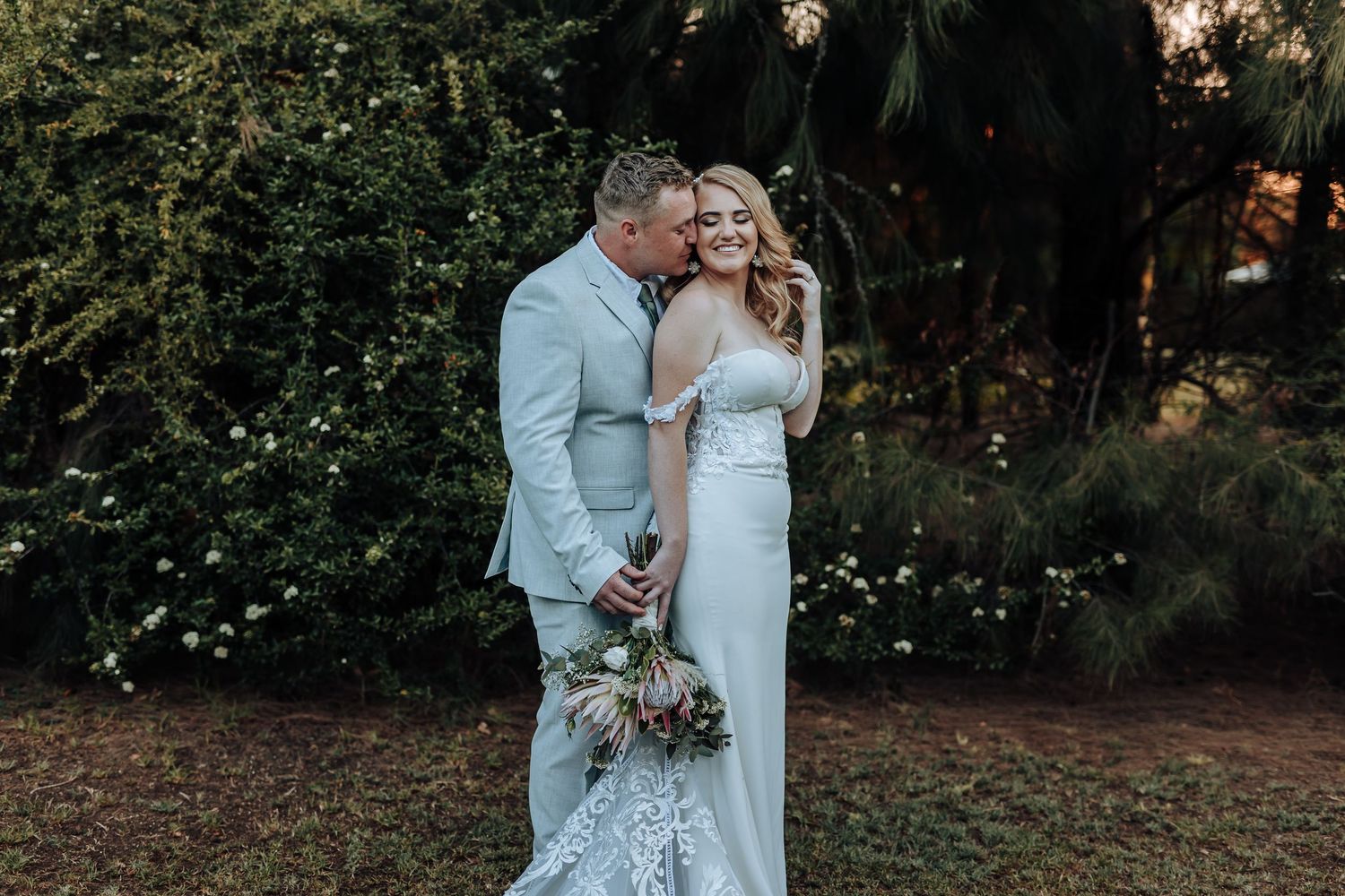 Elegant couple shares intimate moment in garden setting wearing light colored wedding attire.