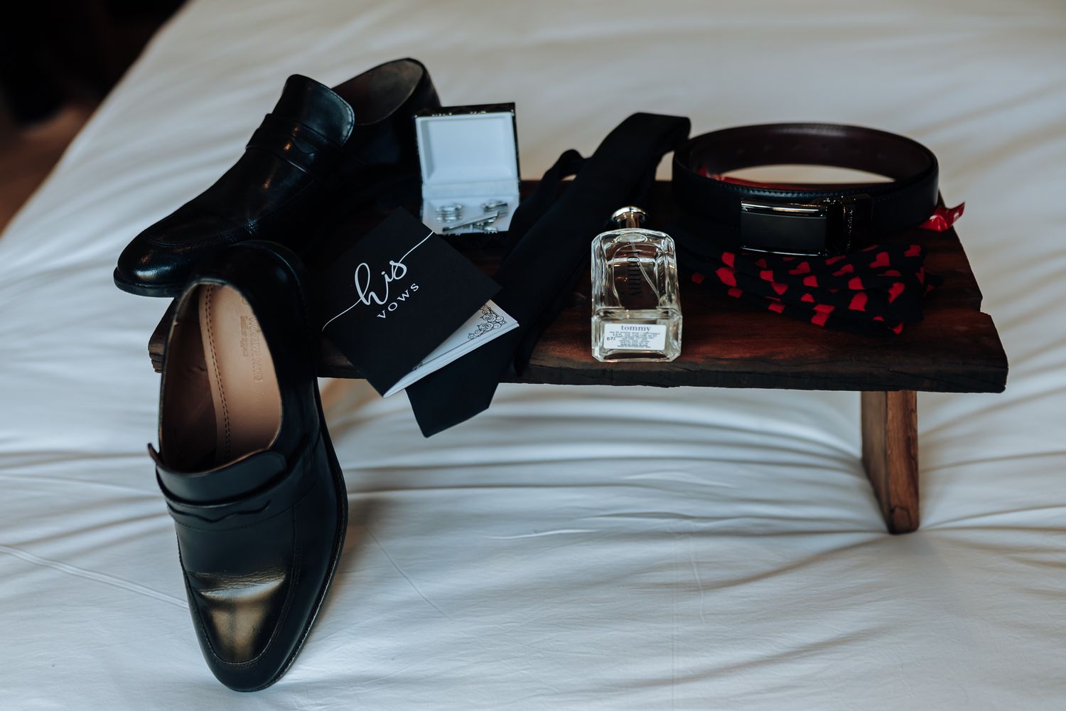 Groom's accessories including black shoes, perfume, and accessories on white bedding.