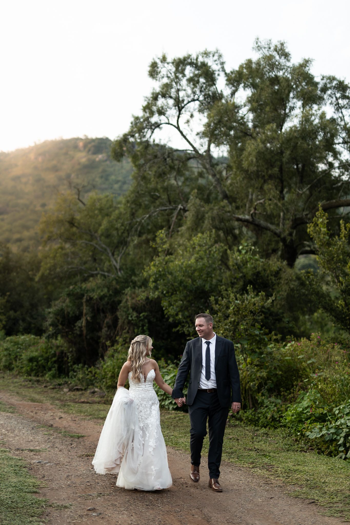 A newlywed couple walks hand in hand along a scenic mountain trail at sunset.