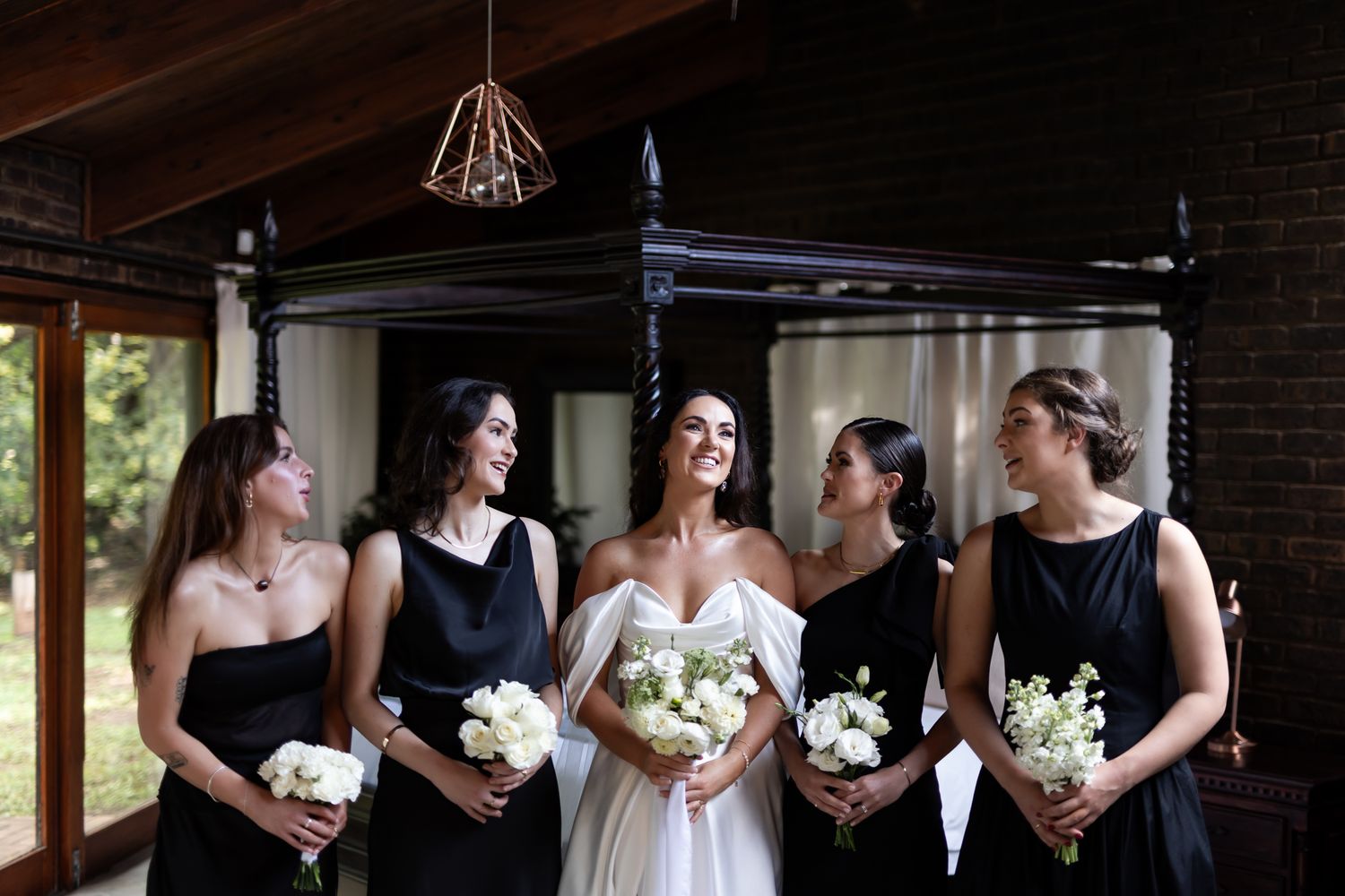 Wedding party poses together near a window with bride in white and bridesmaids in black holding bouquets.