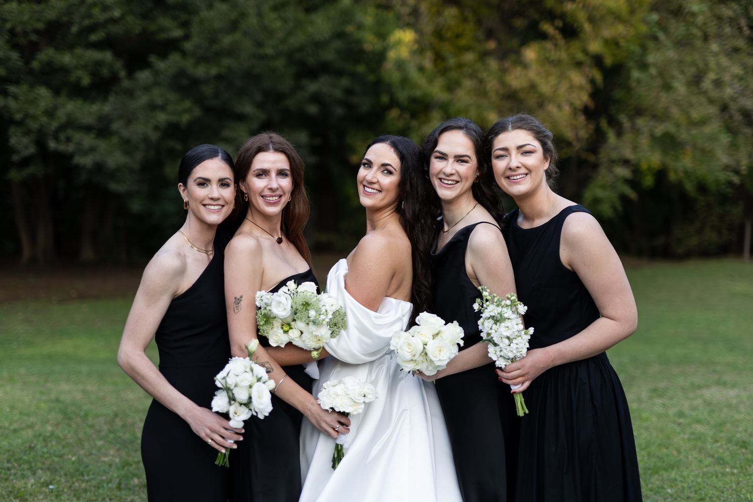 Bridal party in elegant black dresses poses with bouquets against fall background.