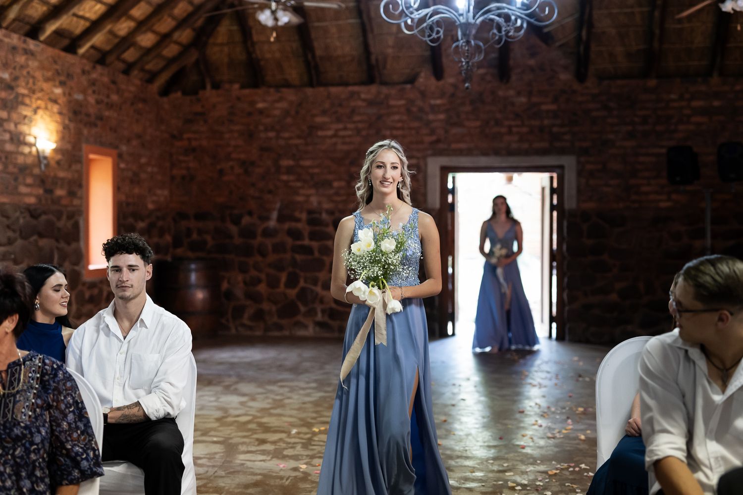 Bridesmaid carrying yellow bouquet stands in flowing blue gown at rustic indoor ceremony venue.