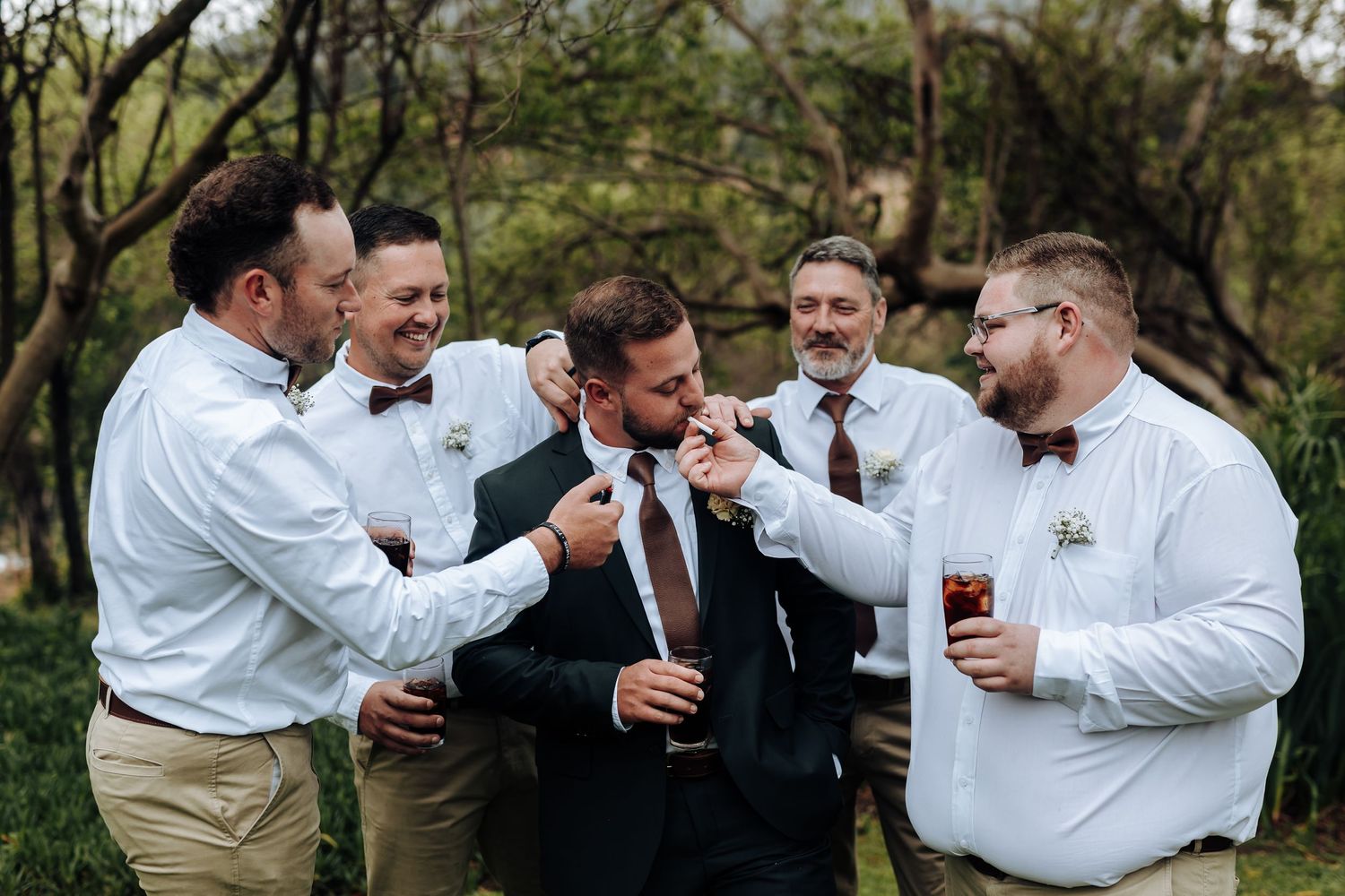 Groomsmen in white shirts and khakis share laughs and drinks during an outdoor wedding celebration.
