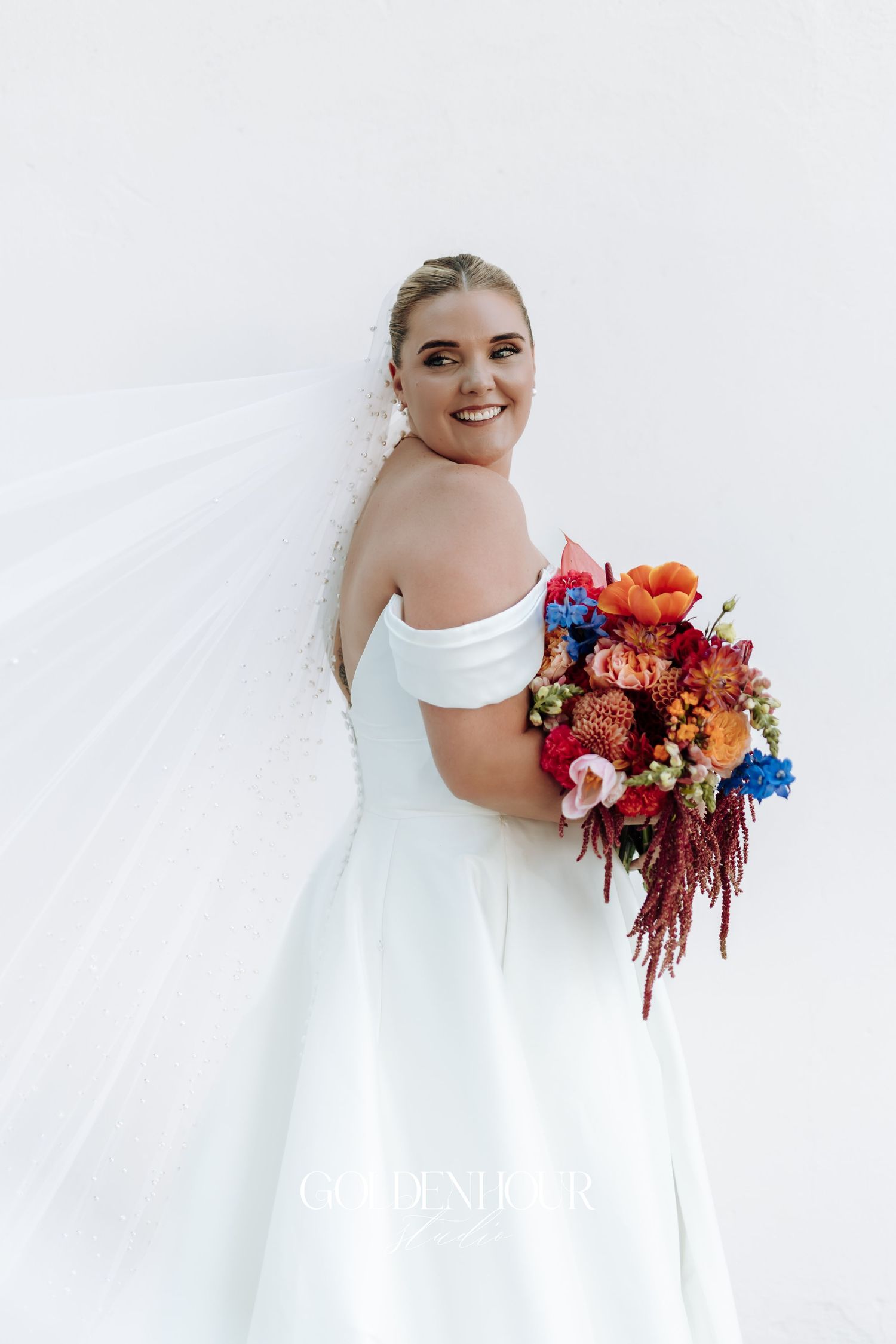 A bride in a white off-shoulder wedding dress holds a vibrant bouquet with orange, blue and red flowers against a light background.