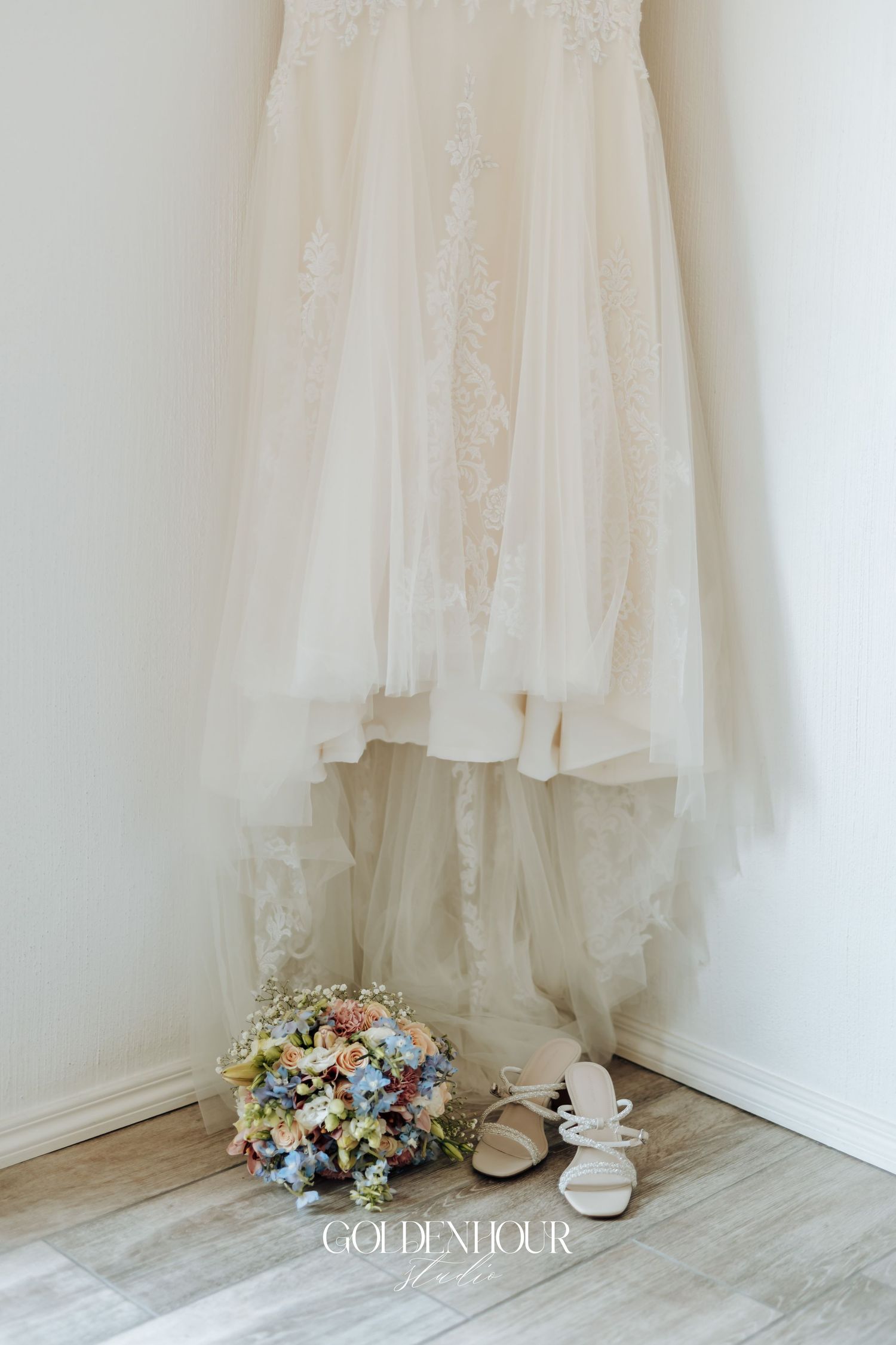 A delicate white wedding dress hangs in a corner with a floral bouquet and shoes arranged below.