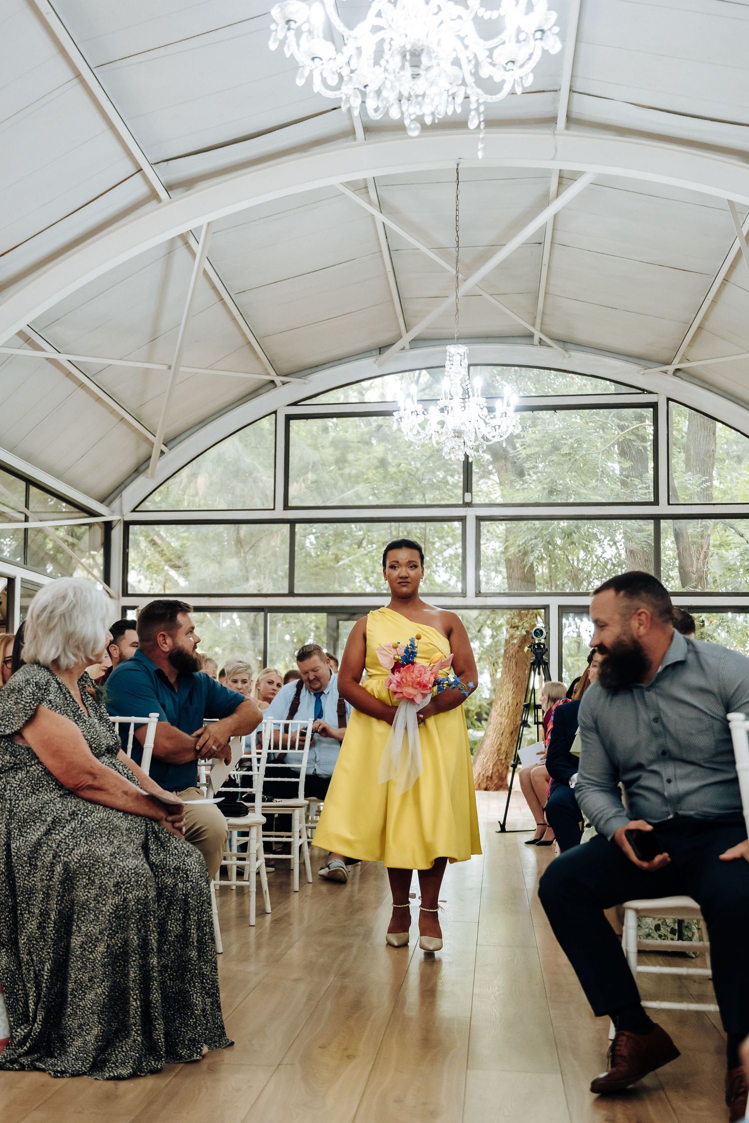 Wedding party member in yellow dress walks down aisle in elegant venue with crystal chandeliers and arched ceiling.