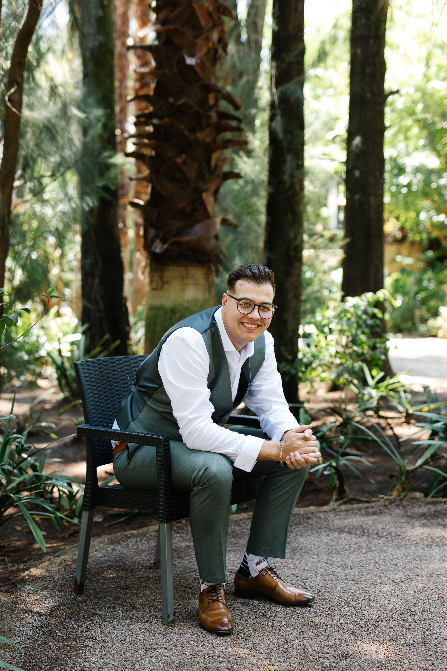 A well-dressed figure in a white shirt and green pants sits on a park bench surrounded by tall trees in a forest setting.