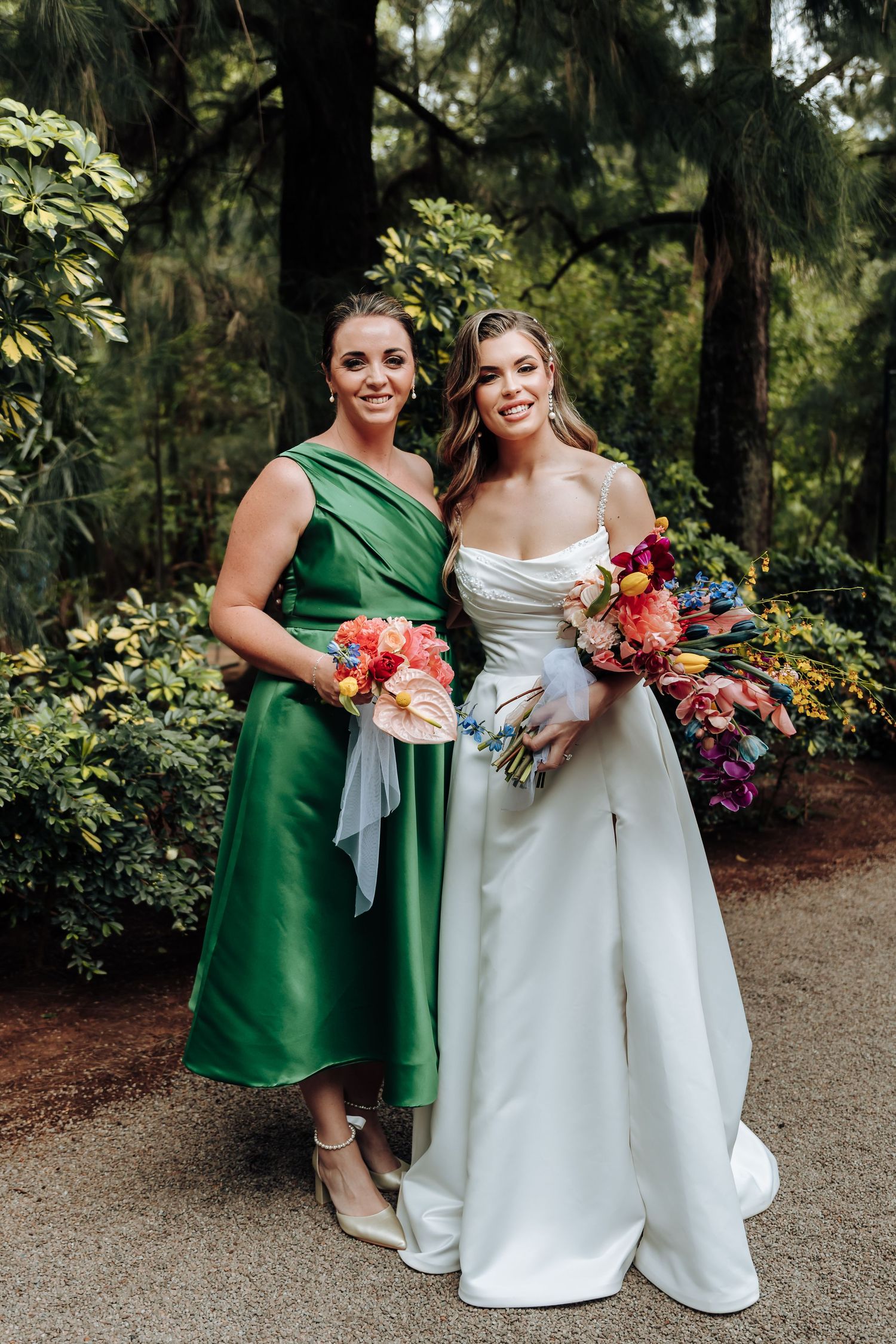A wedding party member in emerald green dress poses with the bride in white gown, both holding vibrant bouquets.