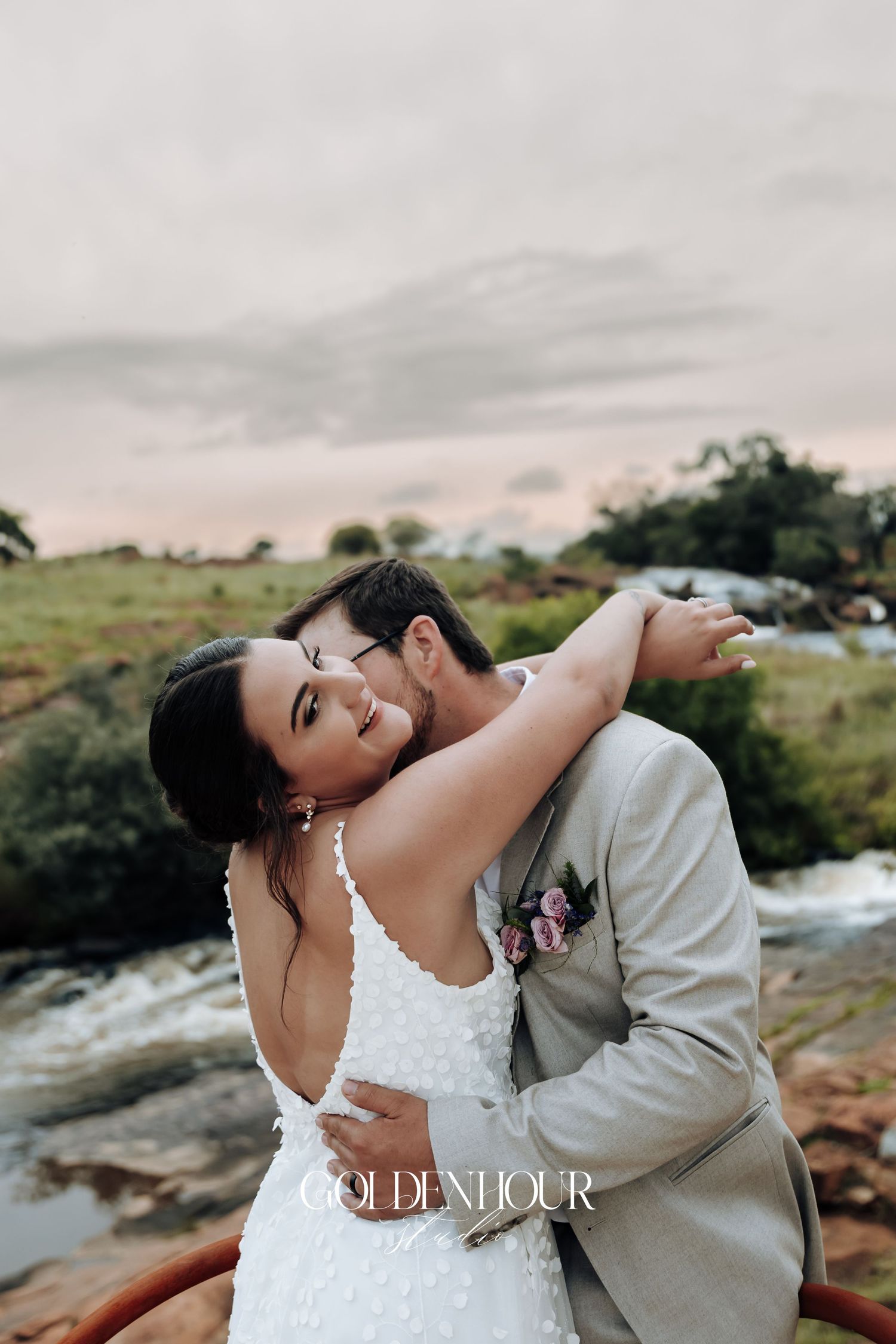Romantic wedding couple embracing in a natural outdoor setting with mountains and water in the background.