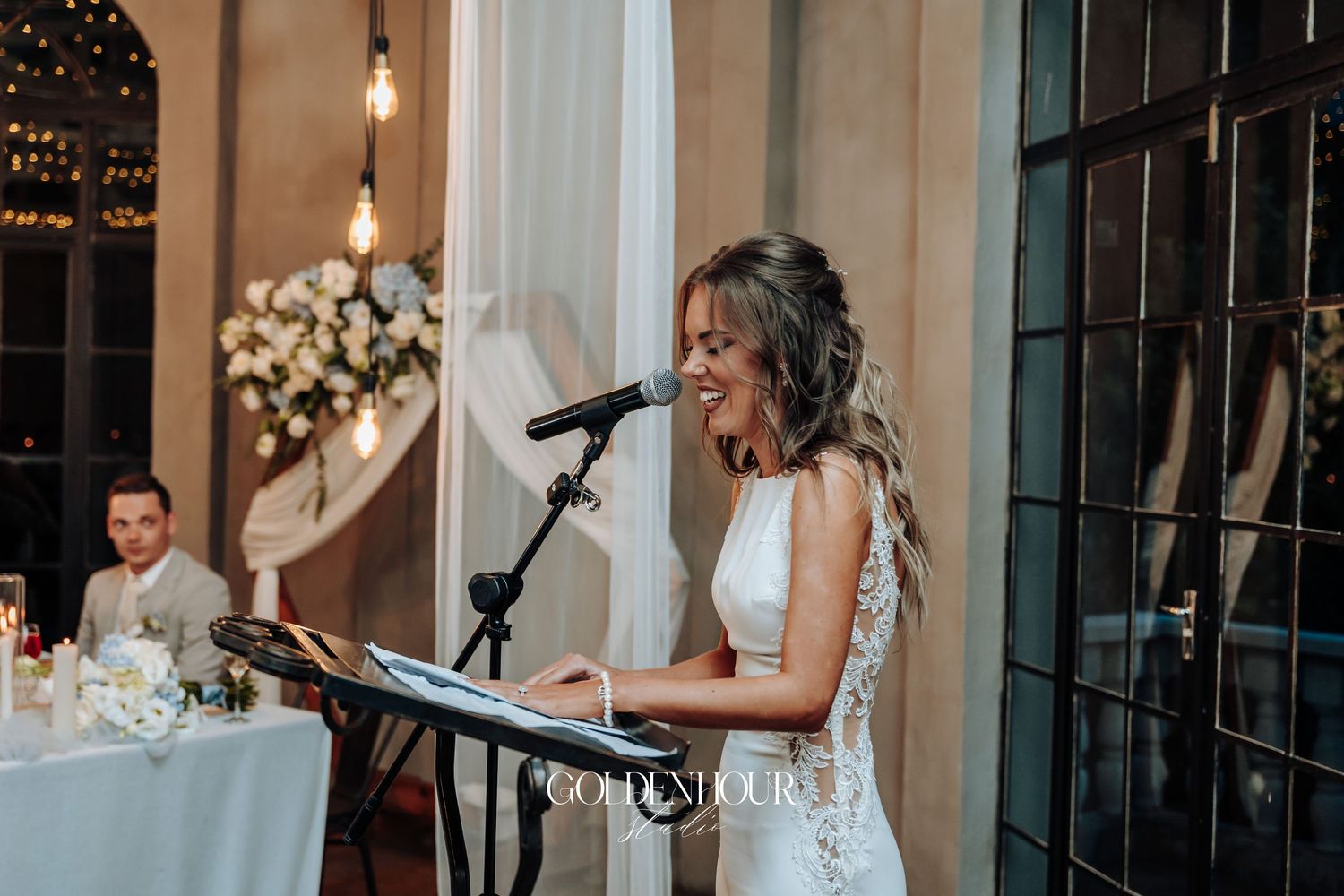 Wedding reception speech being delivered at an elegant indoor venue with floral arrangements.