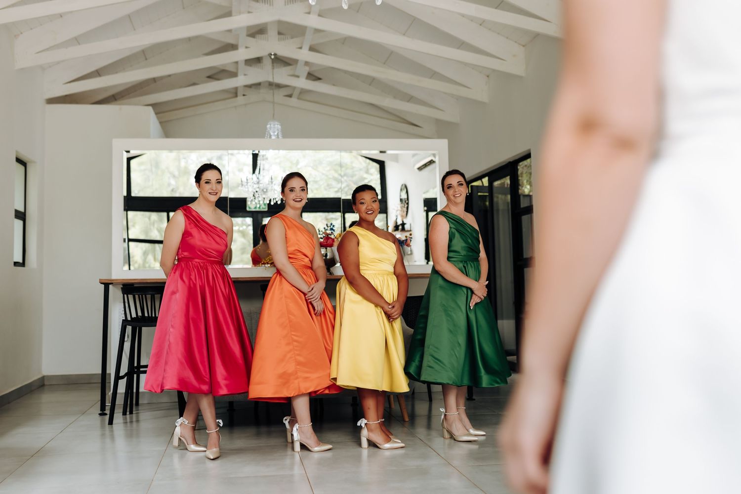 Bridesmaids in vibrant rainbow-colored dresses walk together in a bright indoor space.