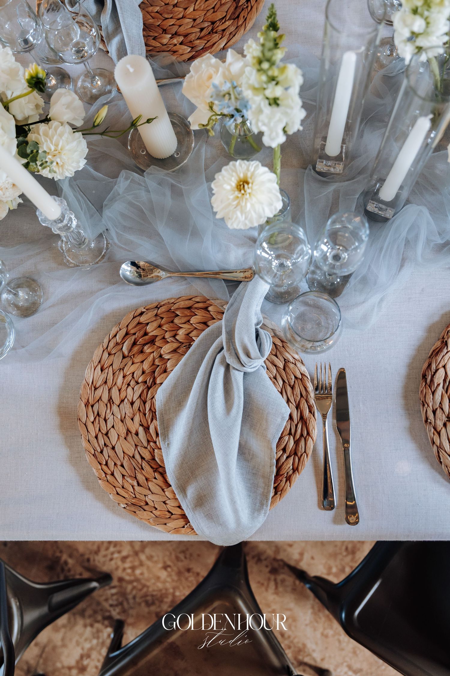 Elegant wedding table setting with white flowers, woven placemats, and crystal glassware on crisp white linens.