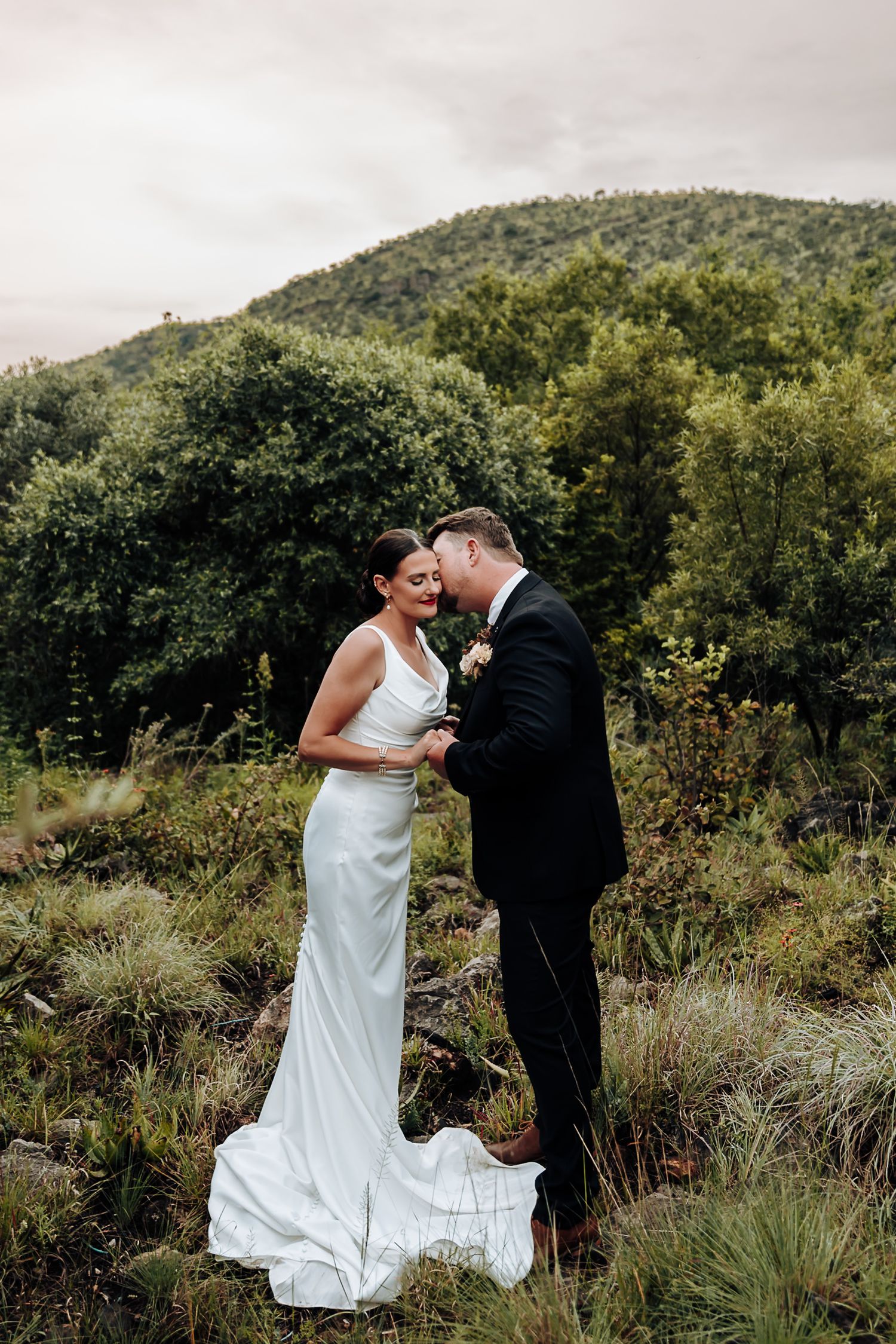 A couple in wedding attire share an intimate moment together against a backdrop of green mountains and foliage.