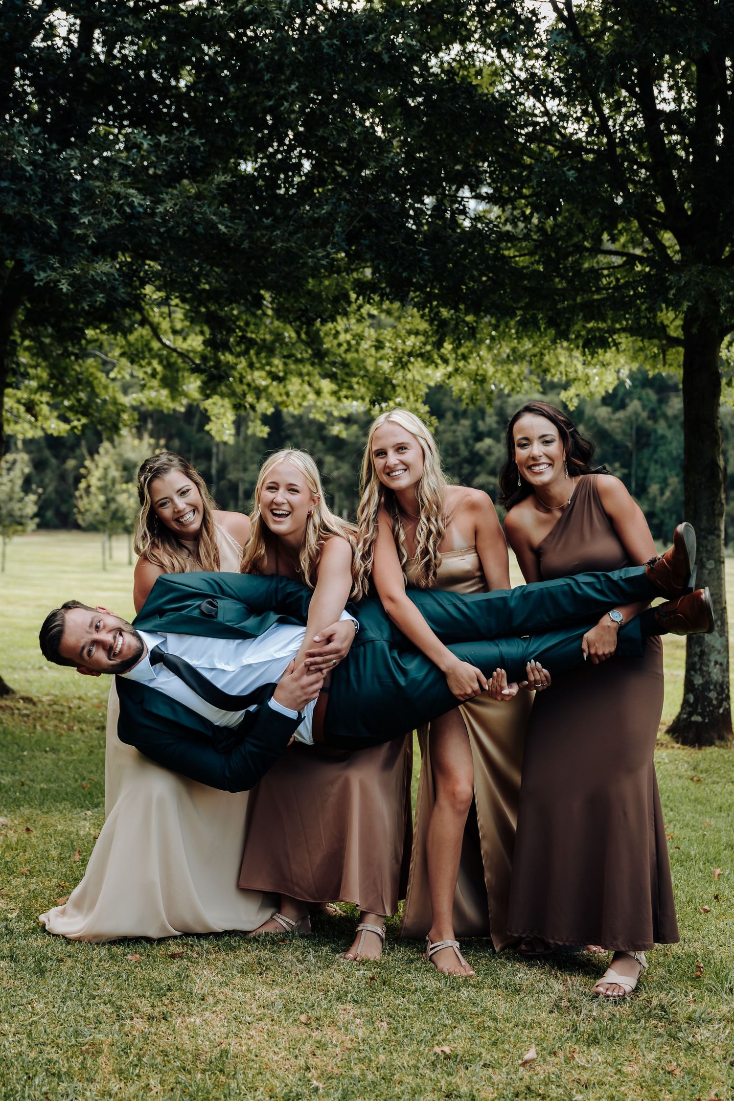 Wedding party members pose playfully on the grass in brown and white dresses for a fun group photo.
