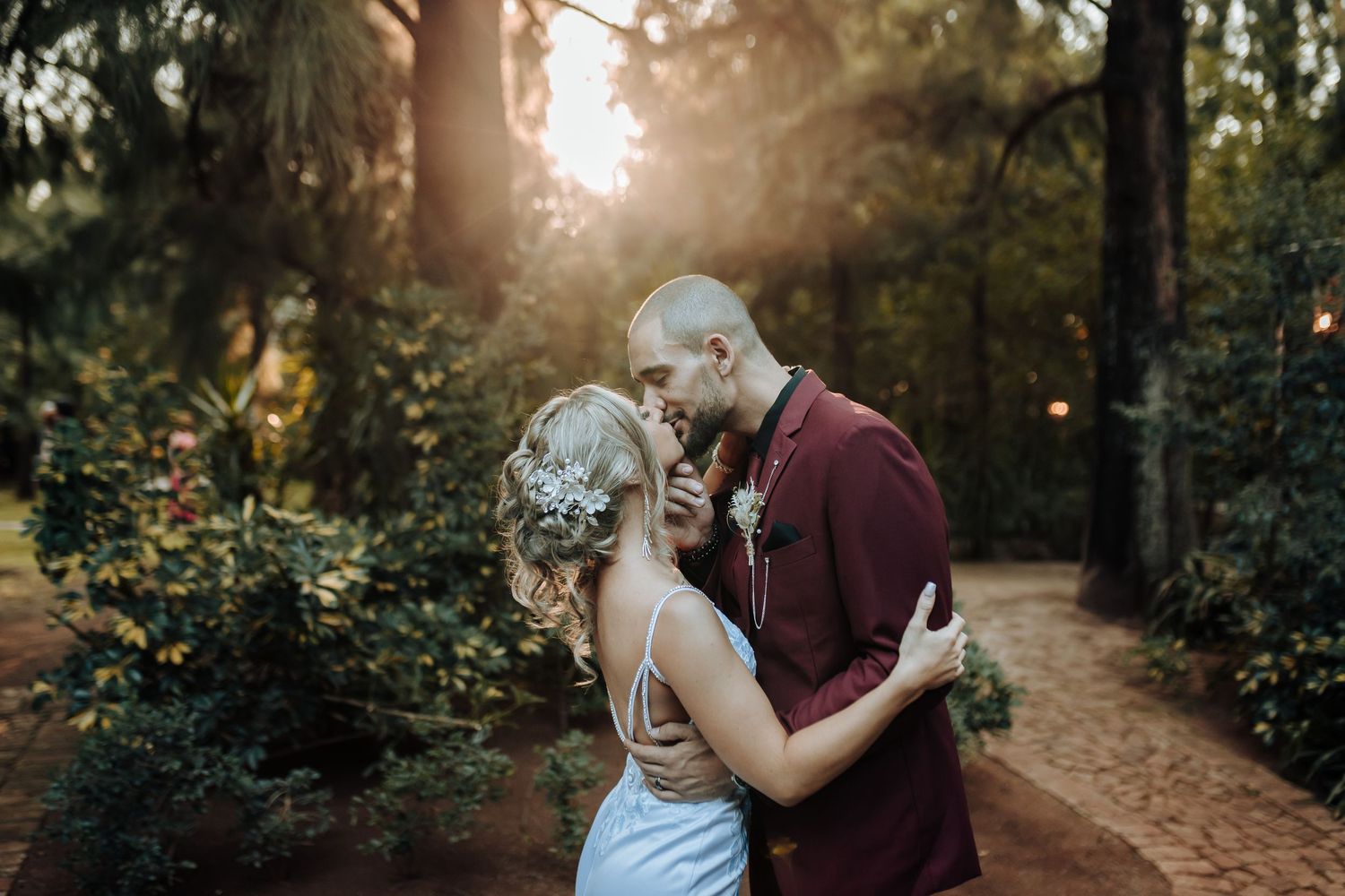 A couple shares a romantic kiss in a sunlit garden path surrounded by lush greenery during their wedding.
