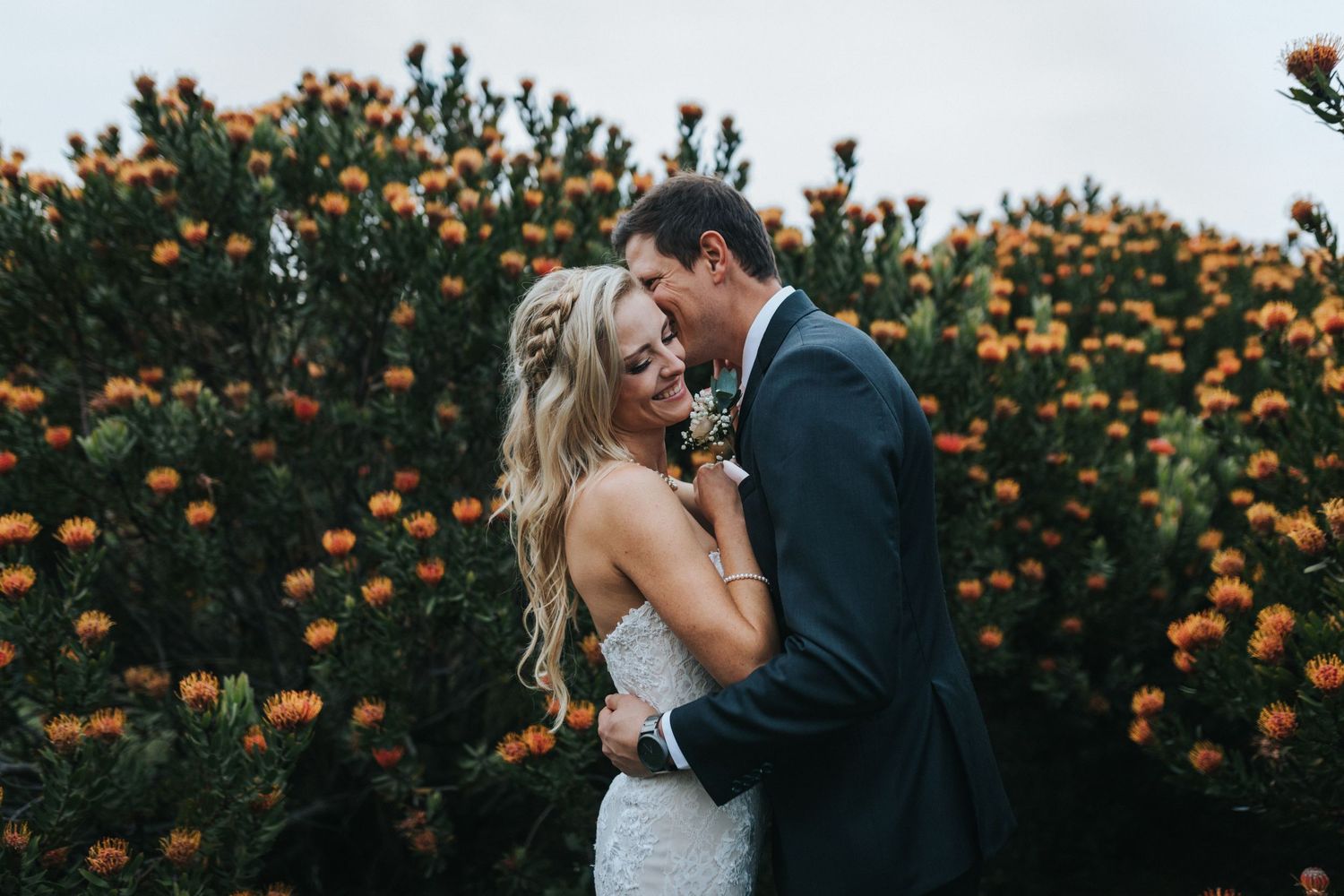 Bride and groom embrace in front of blooming orange flower bushes at their outdoor wedding ceremony.