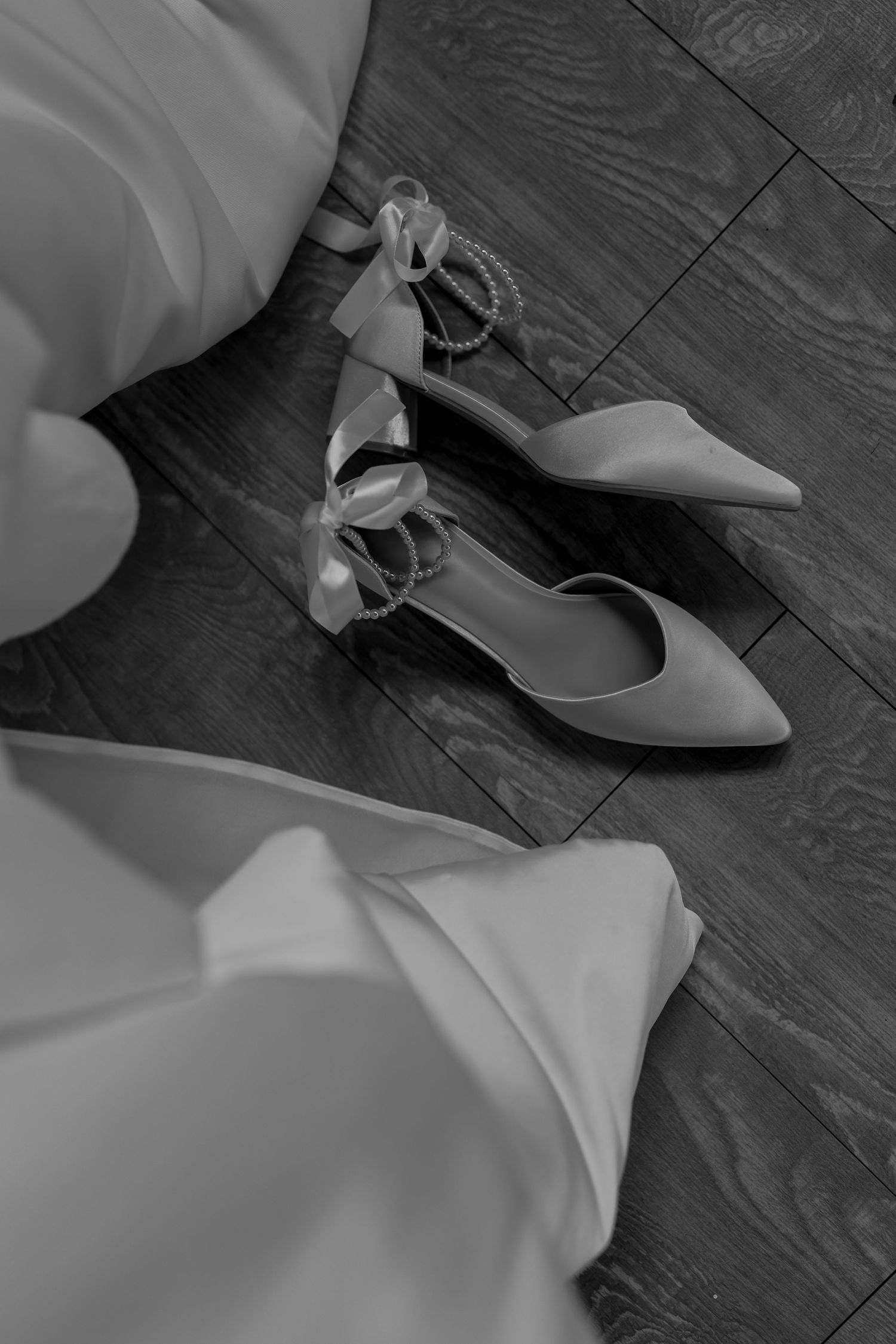 Elegant bridal shoes with ankle straps rest on dark wooden floor beside flowing white fabric.