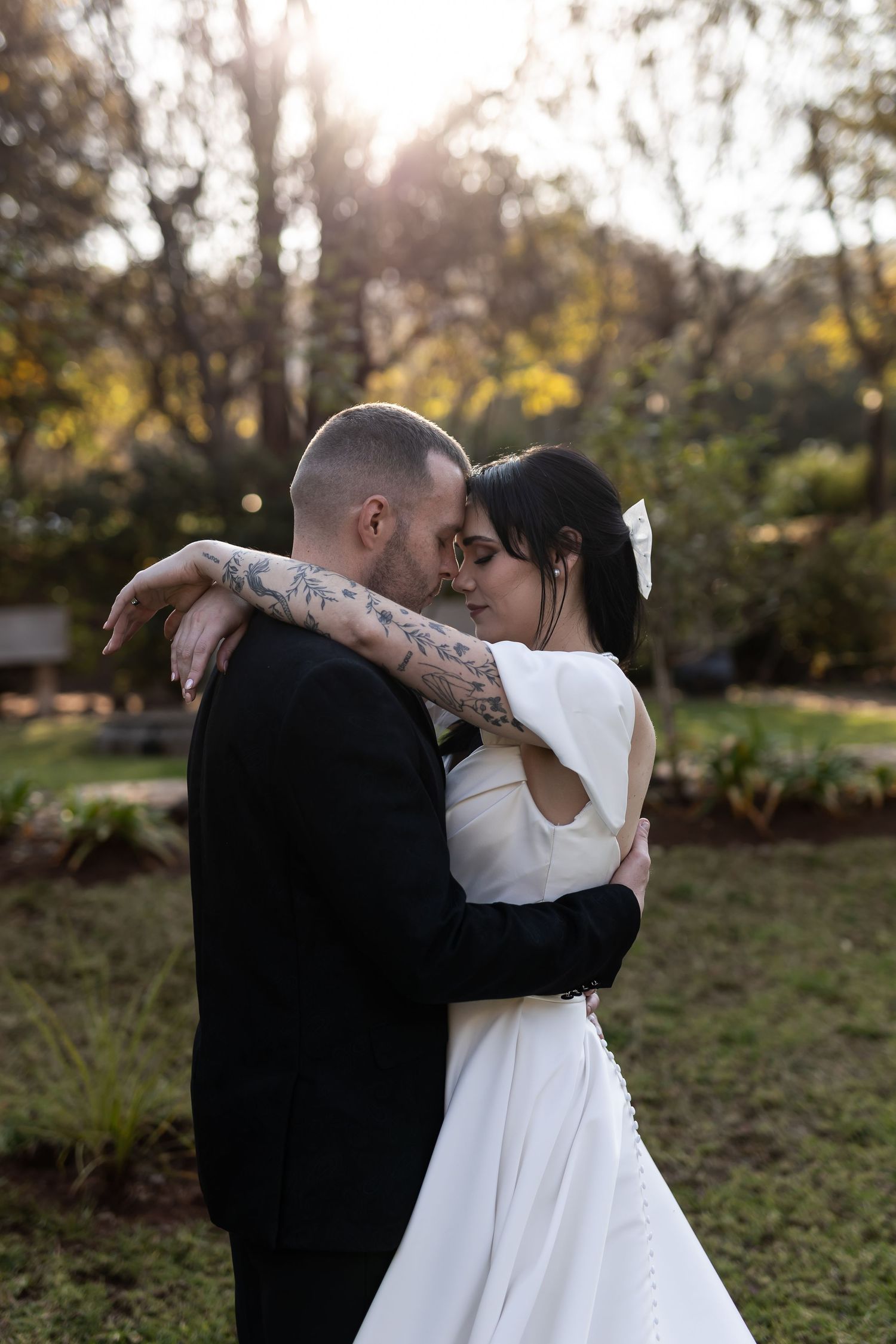 A couple shares an intimate embrace outdoors in a garden setting as sunlight streams through autumn trees.