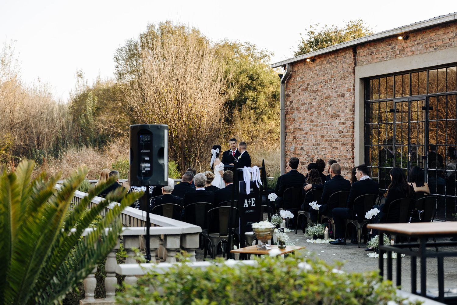 People gather for an outdoor event at a rustic venue with stone walls and patio seating as musicians perform on a black piano.