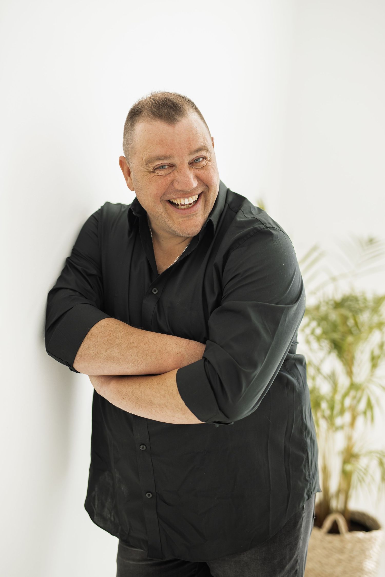 Person in black button-up shirt leaning against white wall with potted plant, smiling warmly in casual pose.