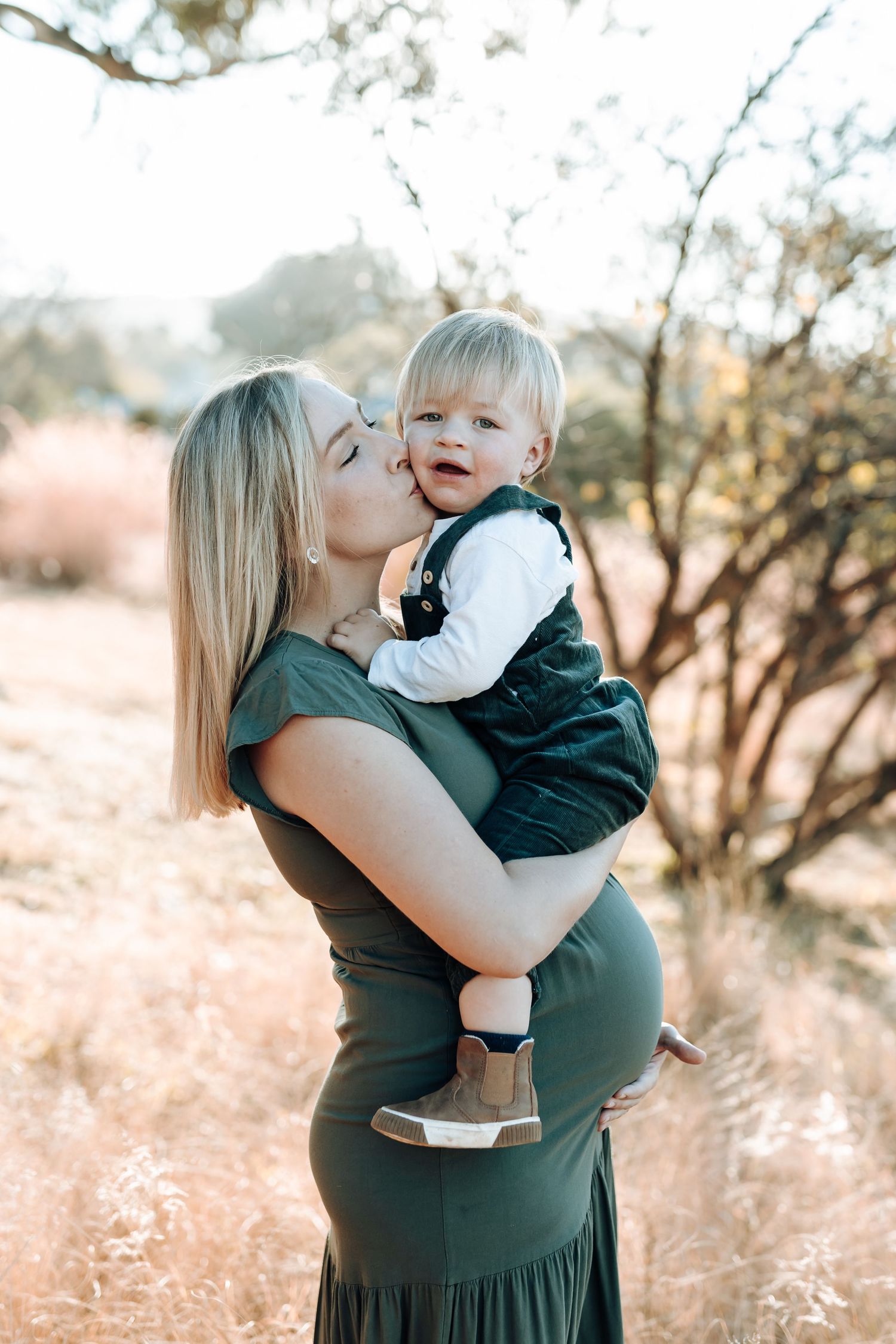 A tender outdoor portrait of a parent and toddler embracing in a sunlit natural setting wearing casual attire.