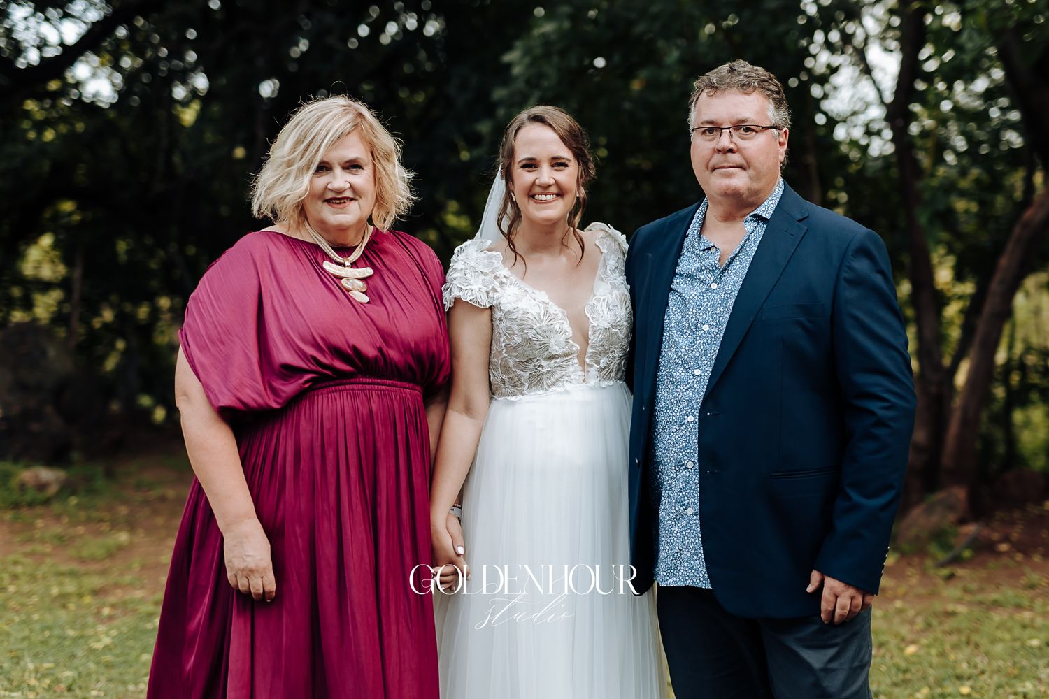 Bride in lace wedding gown posing with parents in burgundy dress and navy suit outdoors.