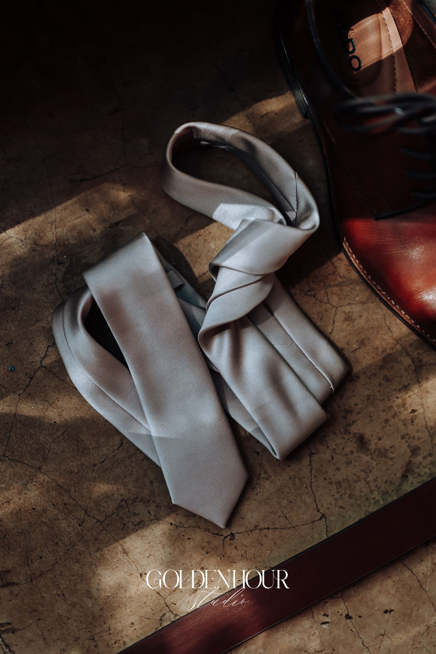Silver tie and brown leather shoes arranged on a textured surface, wedding groom accessories detail shot.
