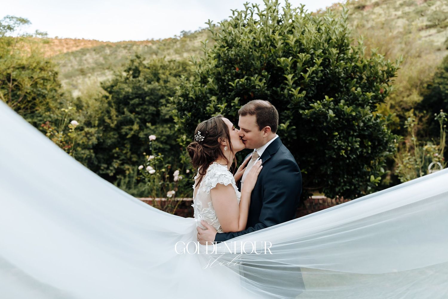 Bride and groom sharing romantic kiss outdoors with flowing veil billowing in the foreground.