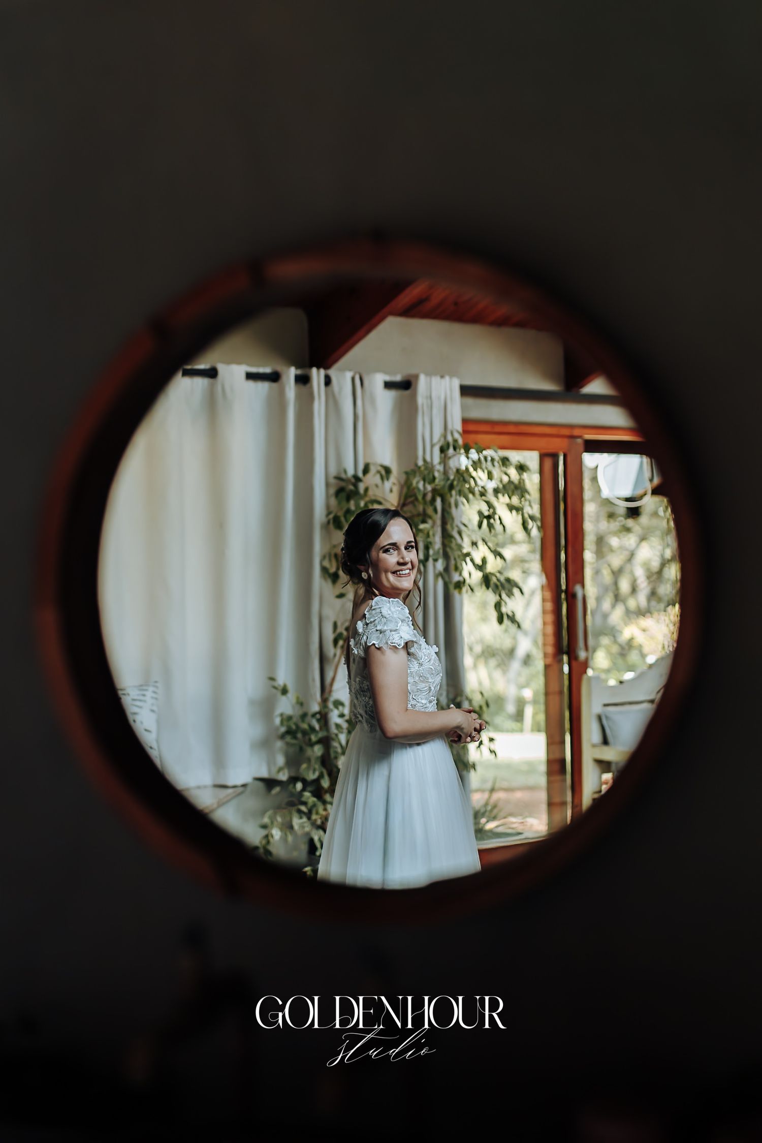 Bride in lace wedding dress smiling, reflected in a round wooden-framed mirror.
