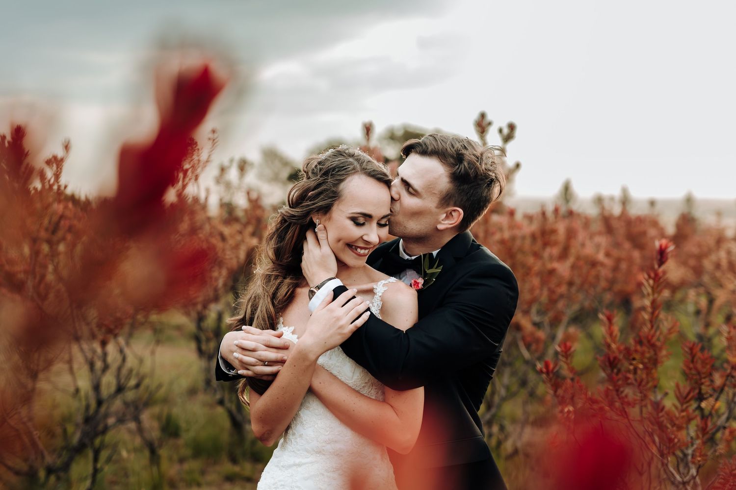 A groom kissing his bride on the cheek while embracing her in a field of red wildflowers.
