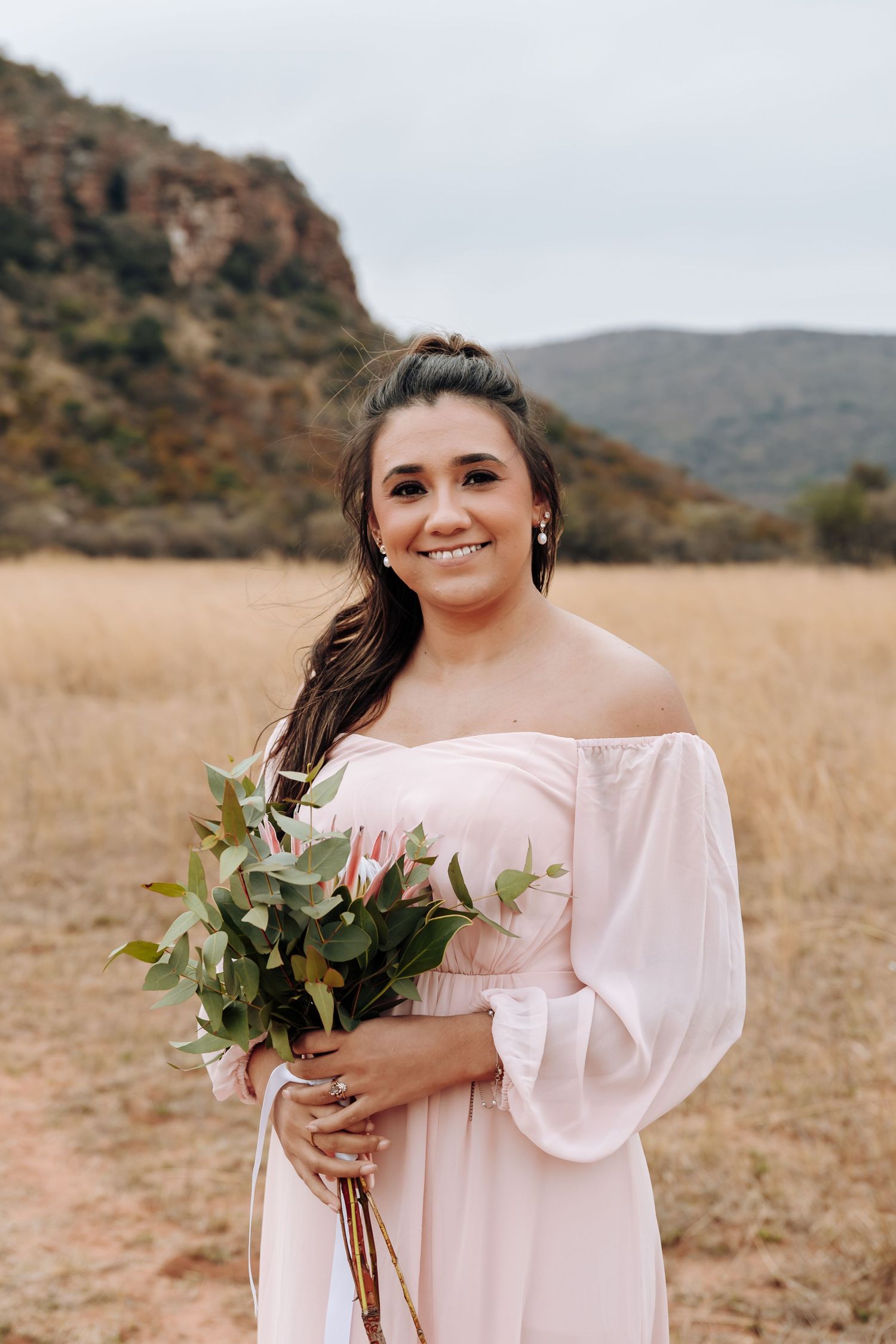 Off-shoulder blush pink dress with blooming bouquet against scenic mountain backdrop in golden field.