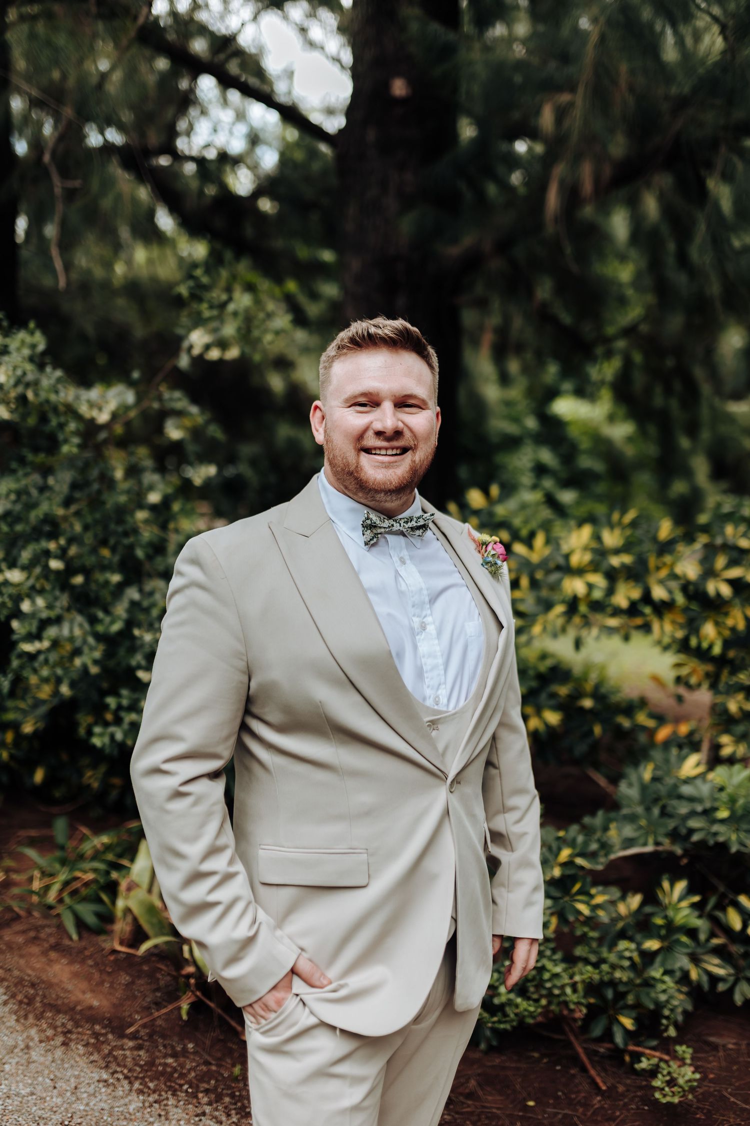 A groom wearing a light beige suit and white bow tie smiles while standing in a lush garden setting.