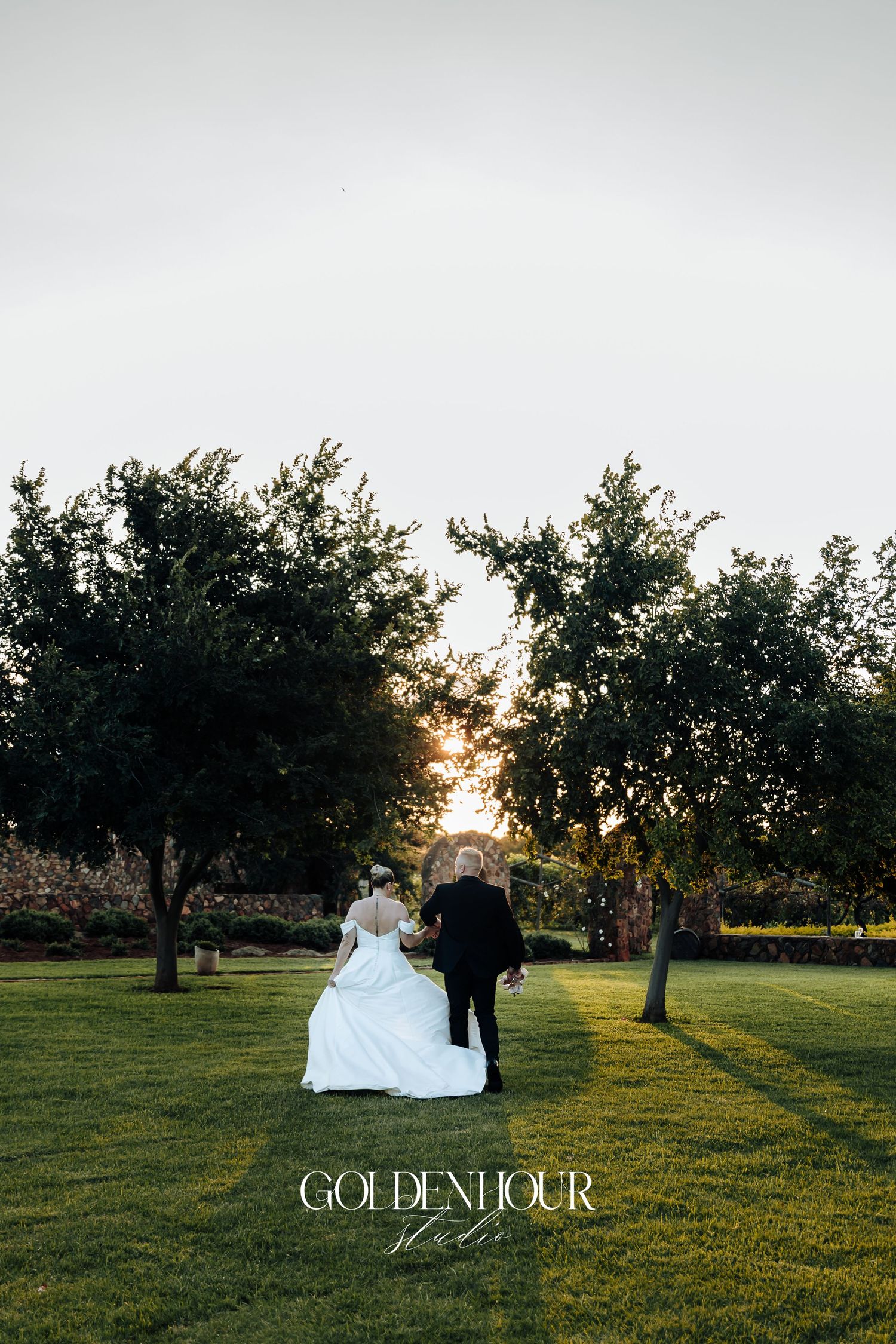 A couple walks across a sunlit lawn during golden hour with trees silhouetted against an overcast sky.