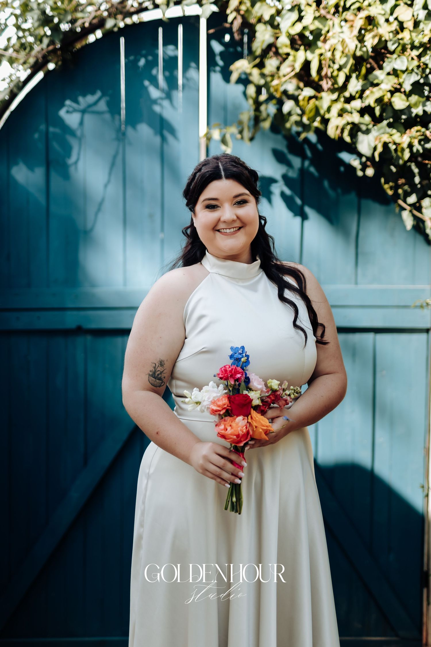 A bride in a cream-colored dress holds a vibrant pink and orange bouquet against a teal fence backdrop.